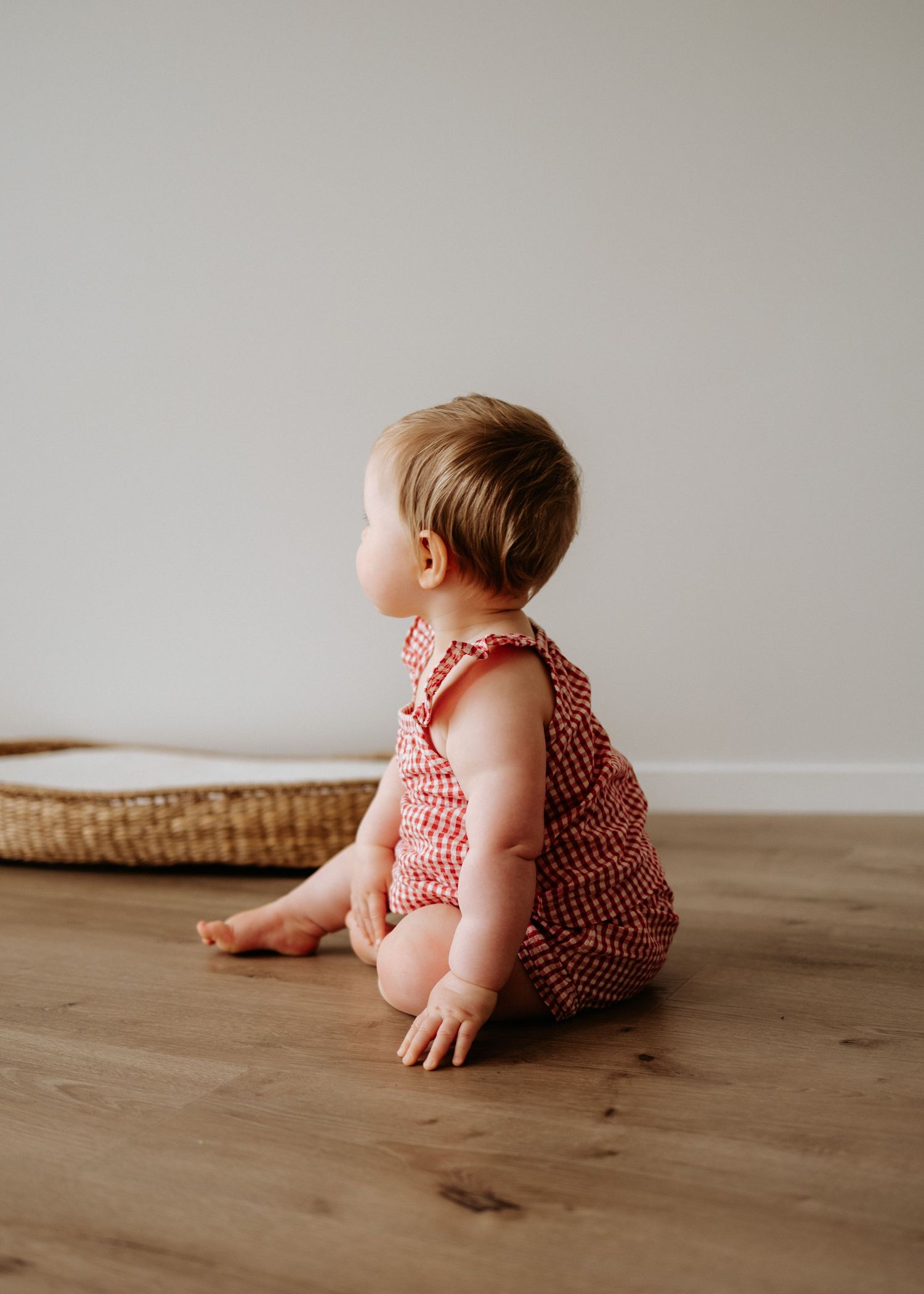 Baby in red floral romper sitting on wooden floor facing away from camera against white wall.