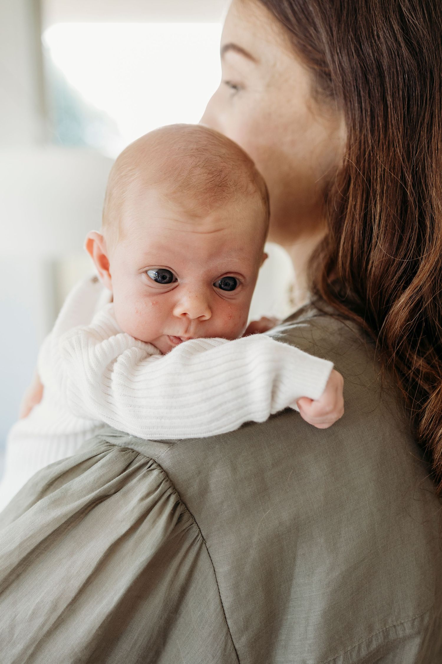 A mother tenderly kisses her newborn baby dressed in white, held over her shoulder against a soft background.