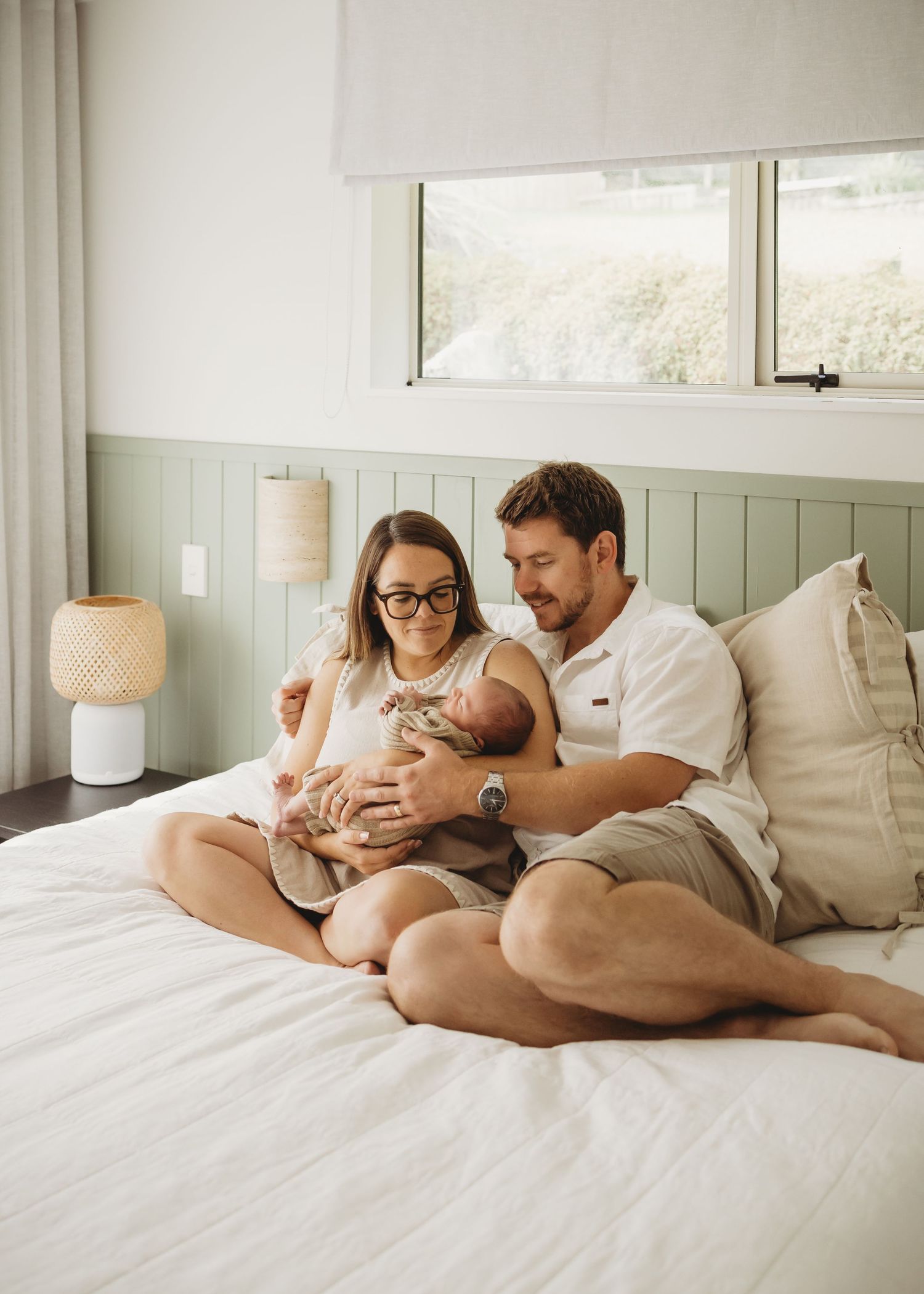 Happy parents sitting on a bed in a bright bedroom admiring their newborn baby.