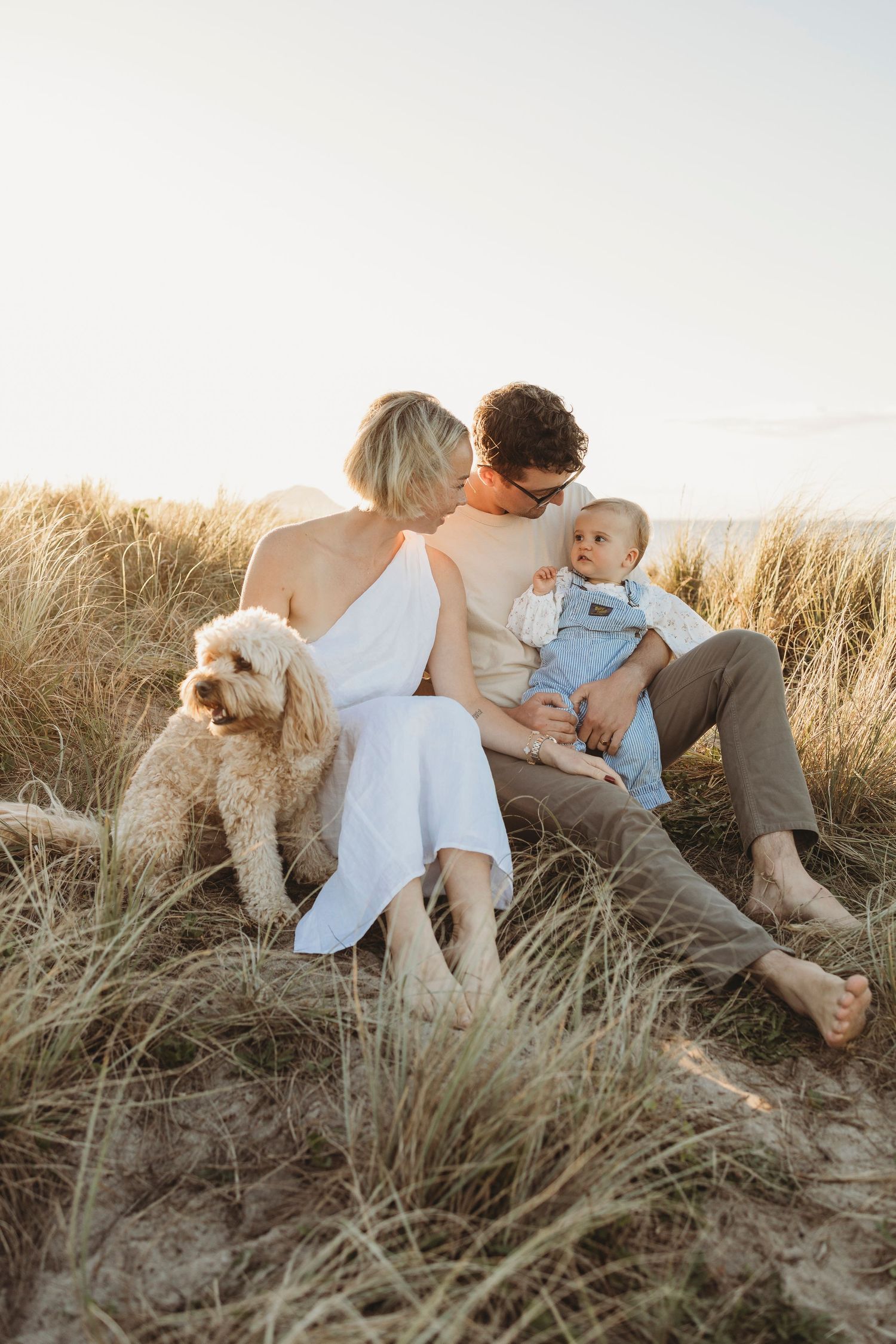 Couple sitting in beach dunes with baby and fluffy white dog during golden hour sunset.