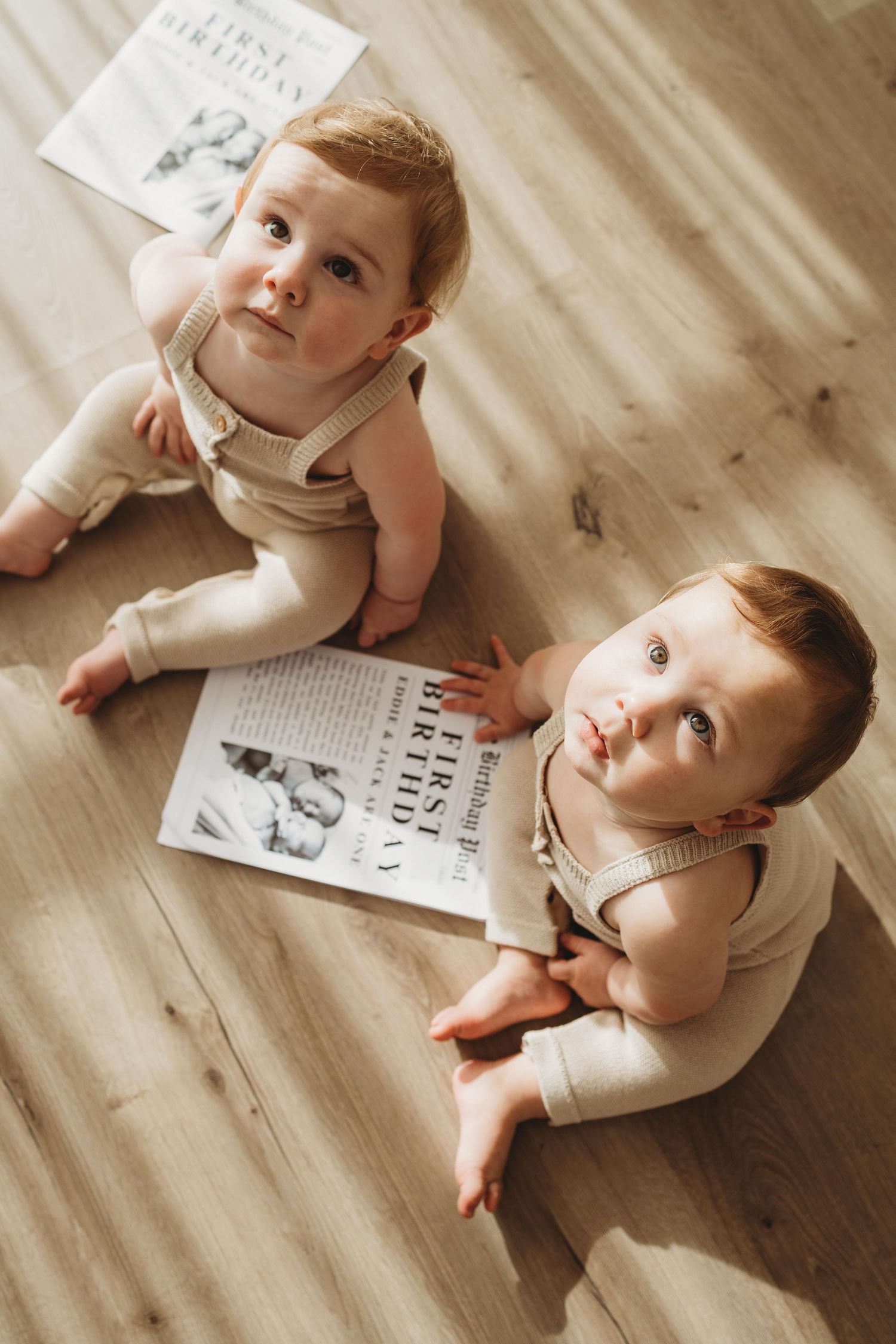 Two red-haired babies in matching beige outfits sitting on wooden floor with first birthday newspaper.