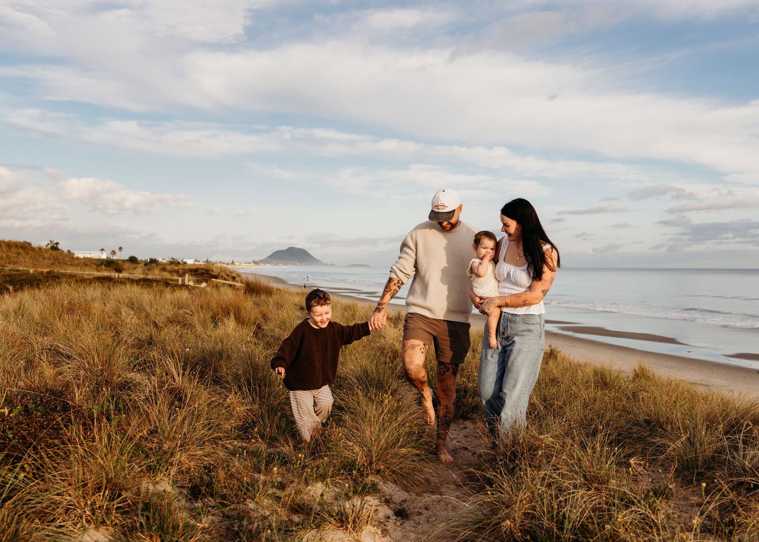 Happy family of four walking along grassy coastal beach path with mountain island in background.