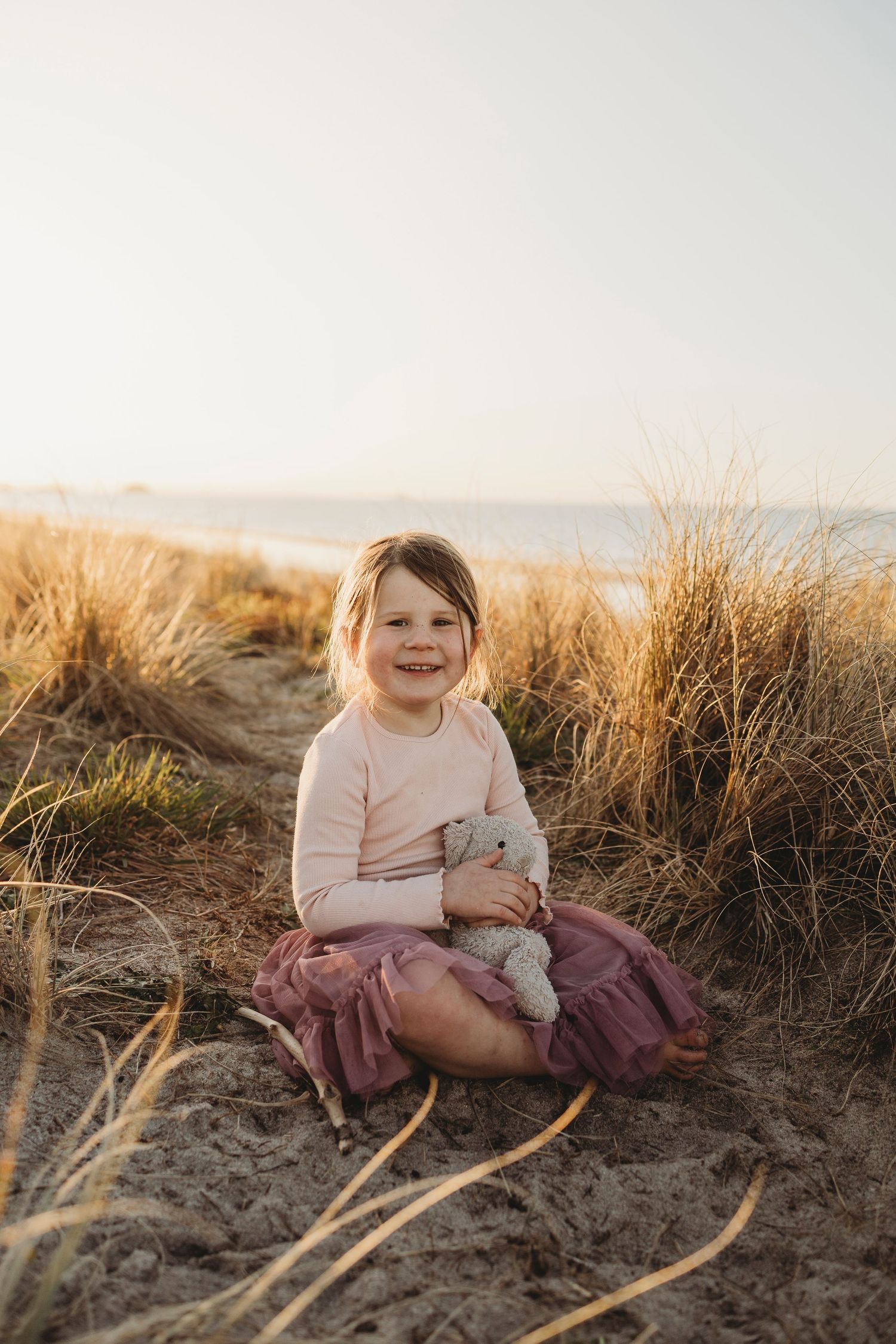 Young girl in pink tutu sits on sandy beach dunes holding stuffed animal, smiling at camera during golden hour.