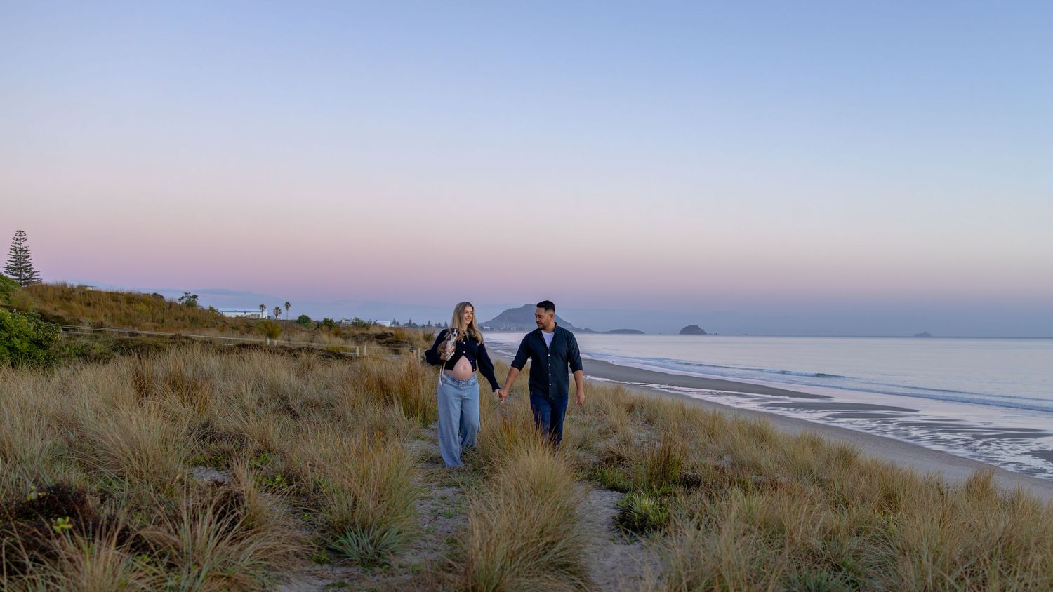 Couple strolling hand in hand through beach grass at twilight with calm ocean and pink sky.