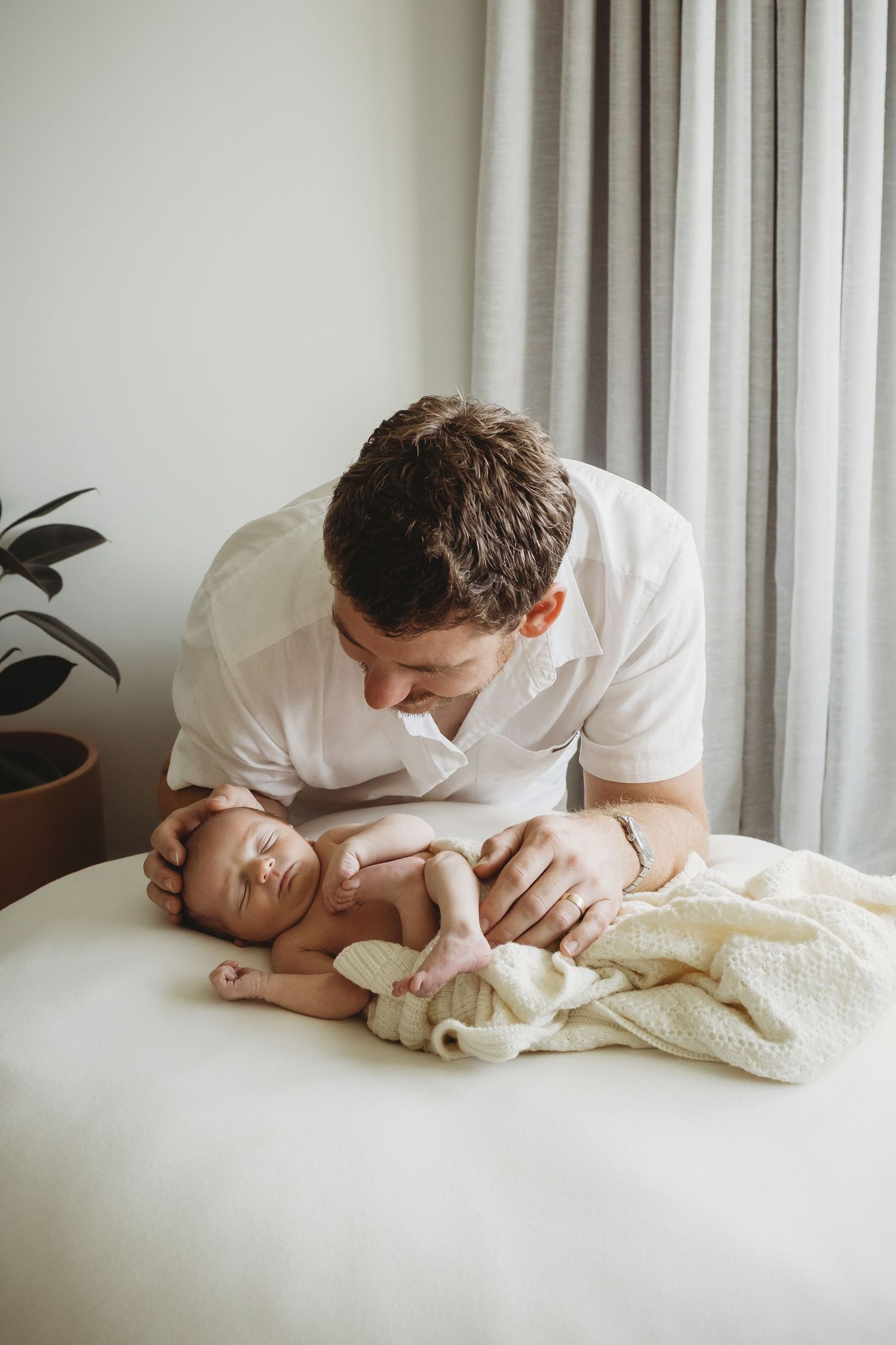 Smiling father leaning over sleeping newborn baby on white surface near window.