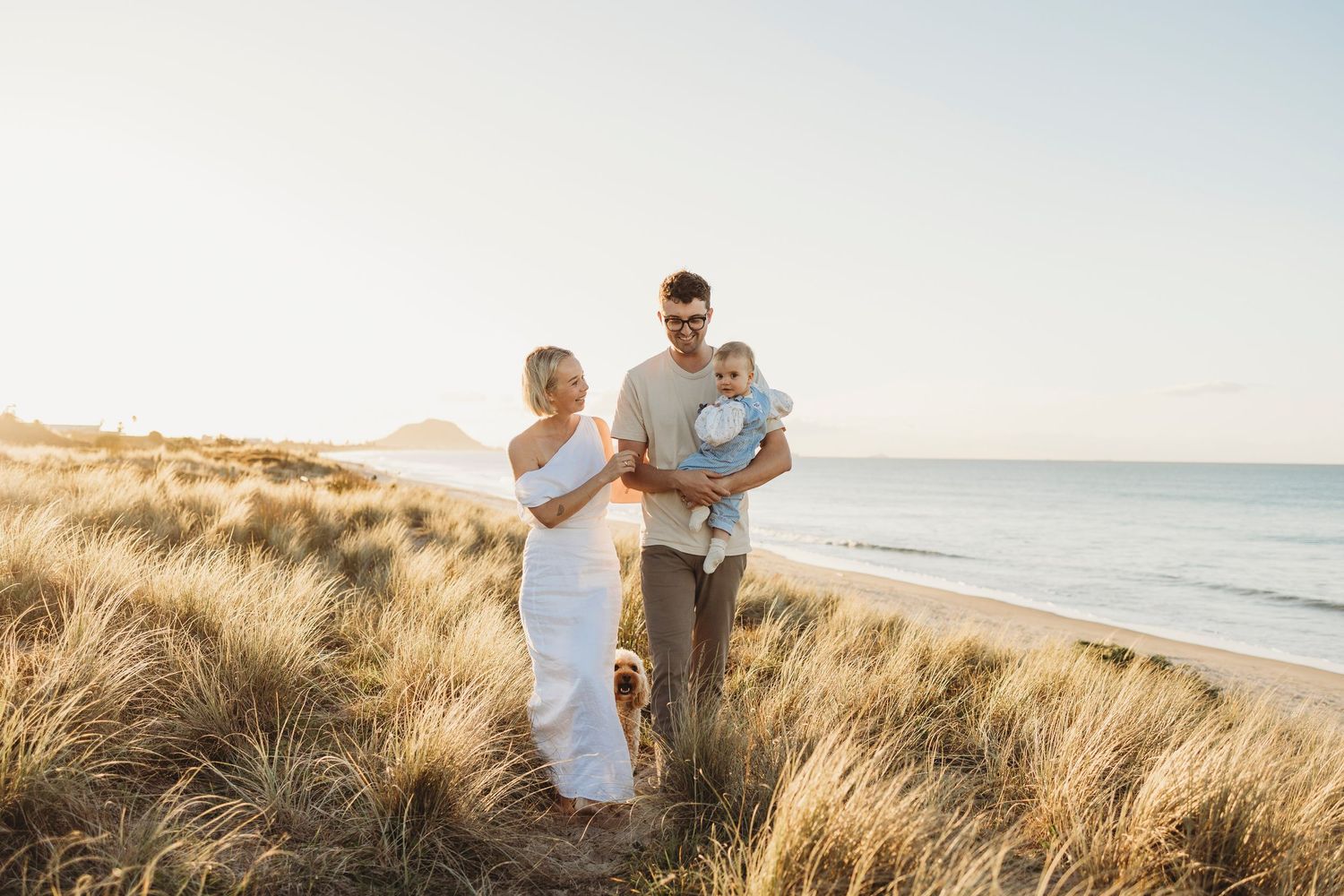Young family of three standing in sunny beach dunes, father holding baby with ocean behind them.