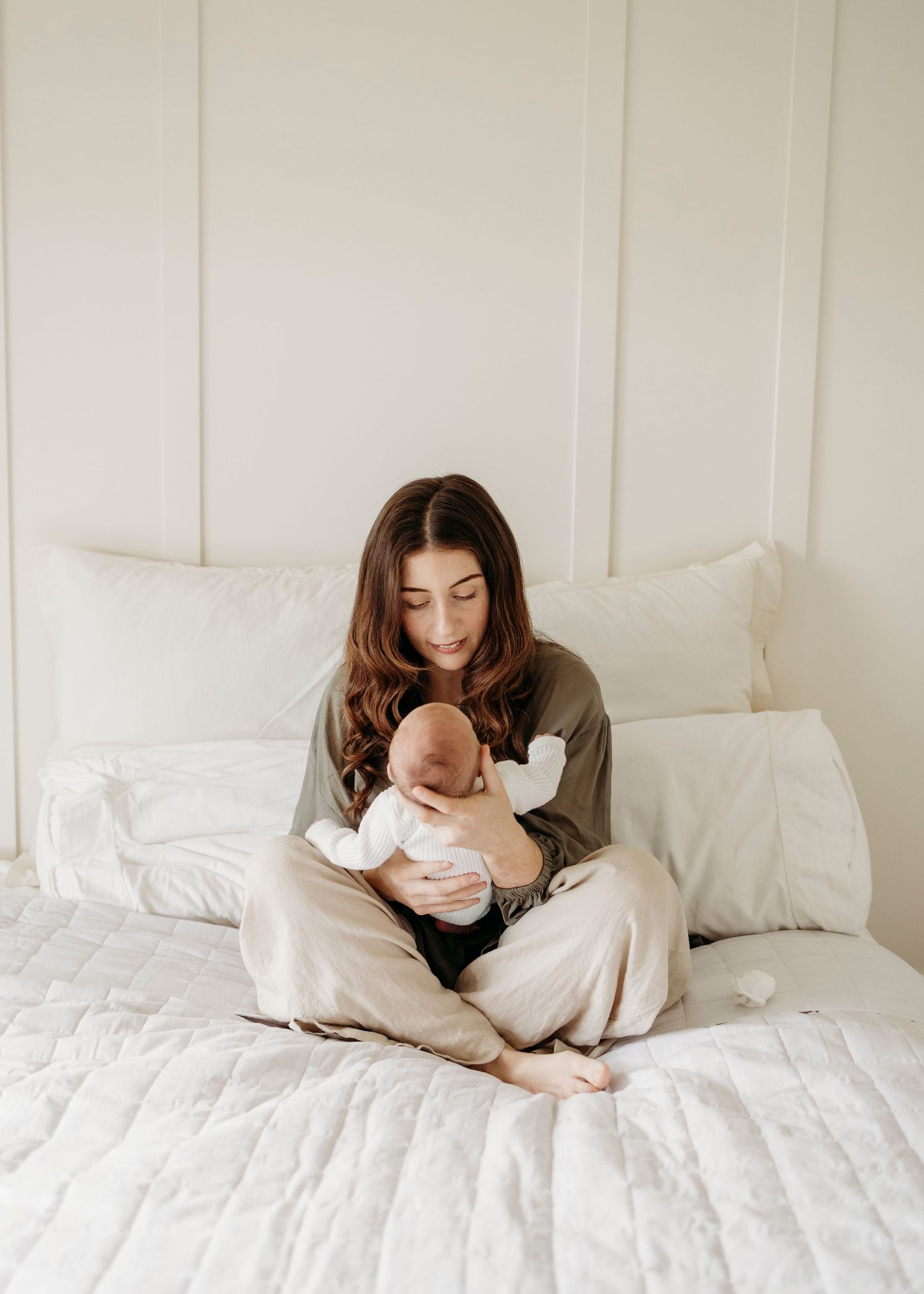 Young brunette mother sitting cross-legged on white bed cradling her newborn baby.