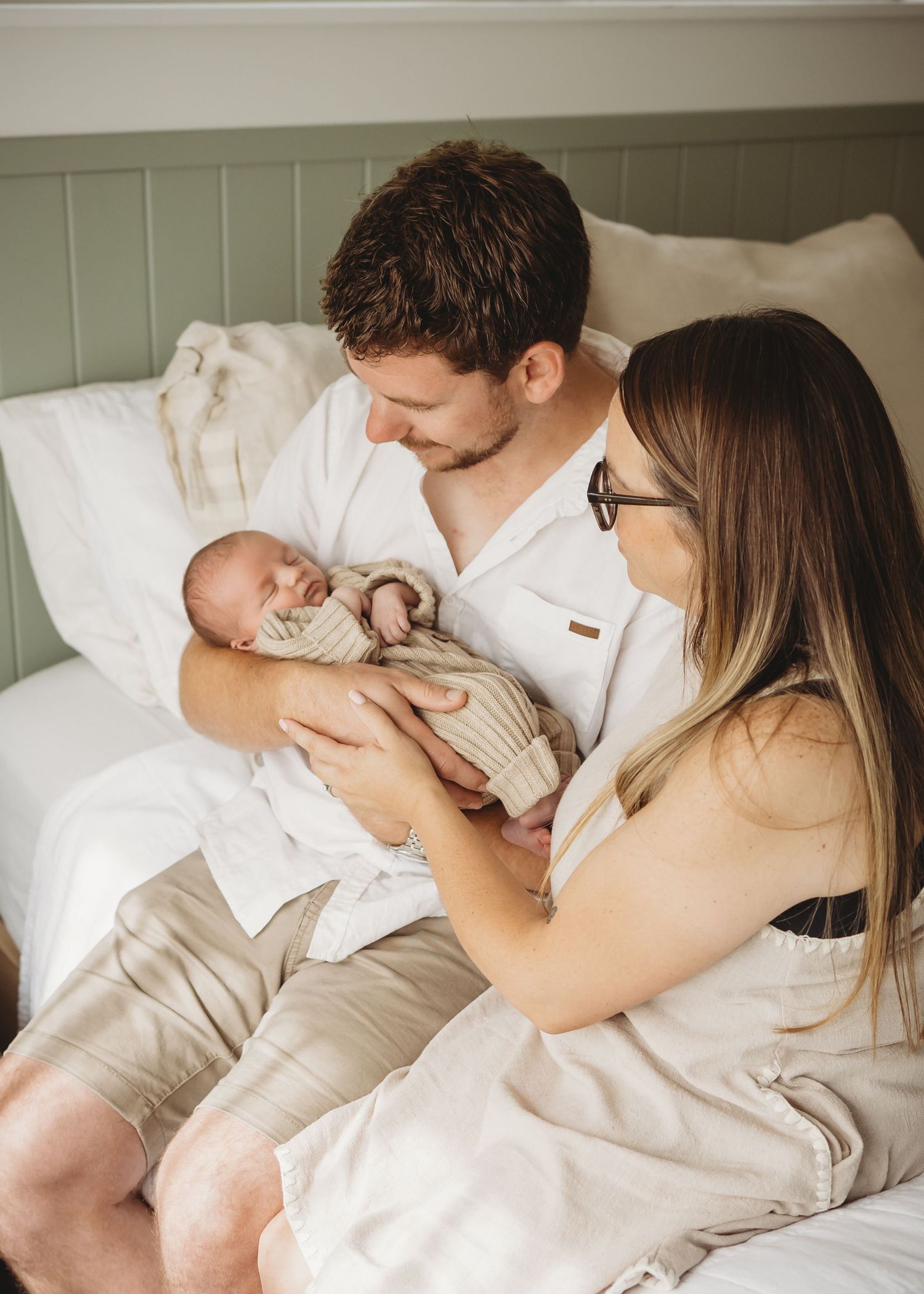 New parents sitting on bed together lovingly gazing at their sleeping newborn baby in knit outfit.