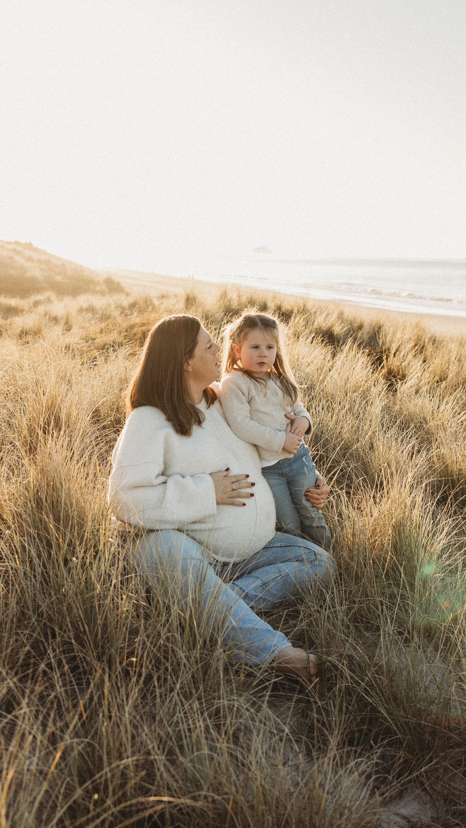 Pregnant woman in white sweater sitting in beach dunes holding young daughter at sunset.
