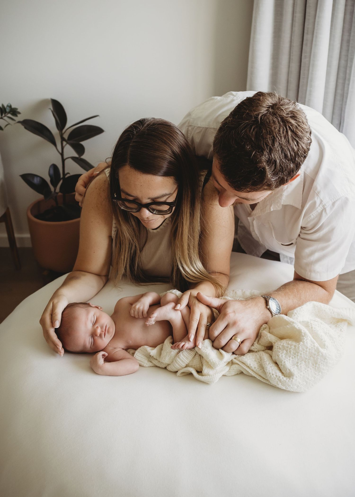 New parents lovingly gazing at their sleeping newborn baby on a white bed.