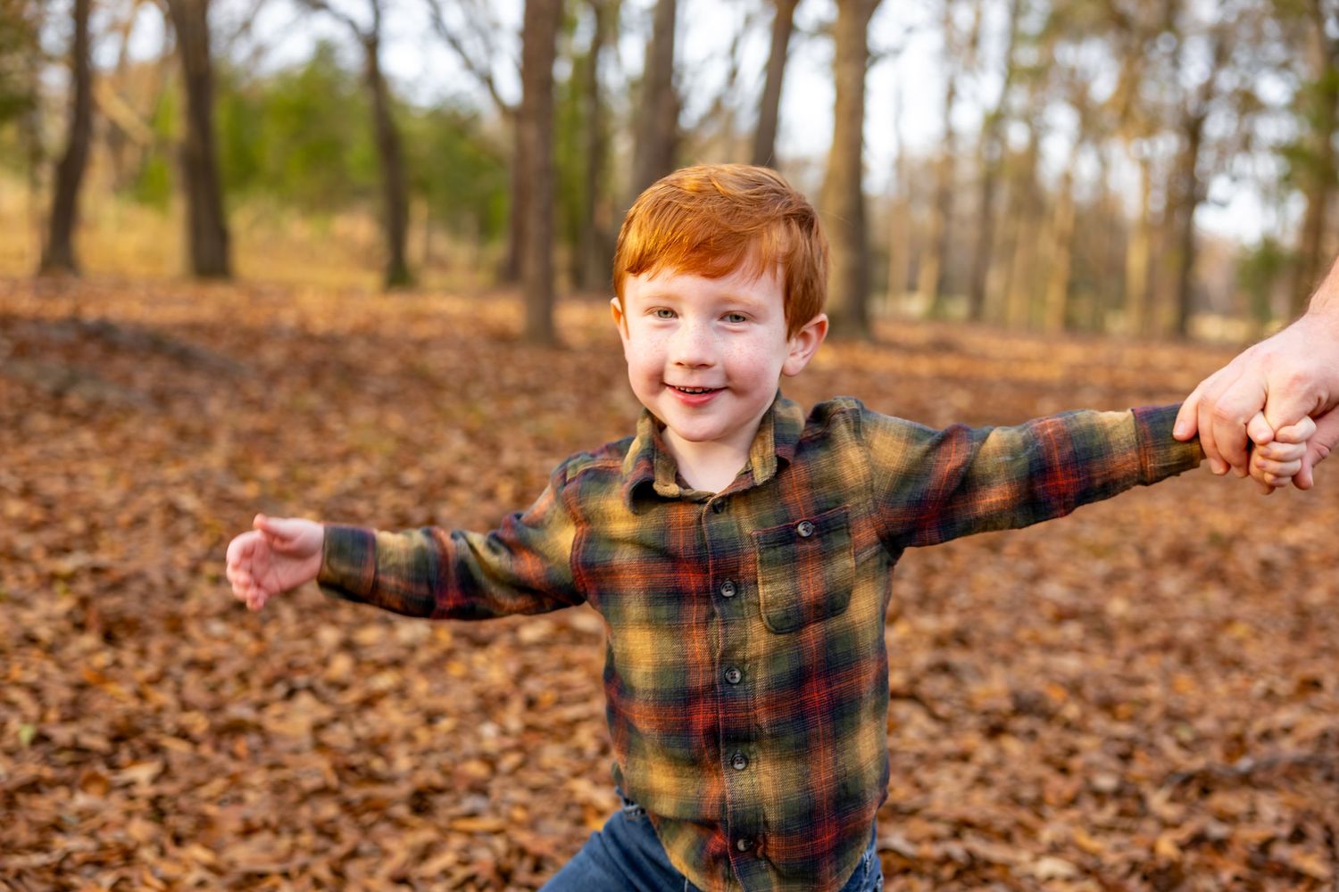 Child in plaid shirt playing joyfully among fallen leaves in a wooded area.