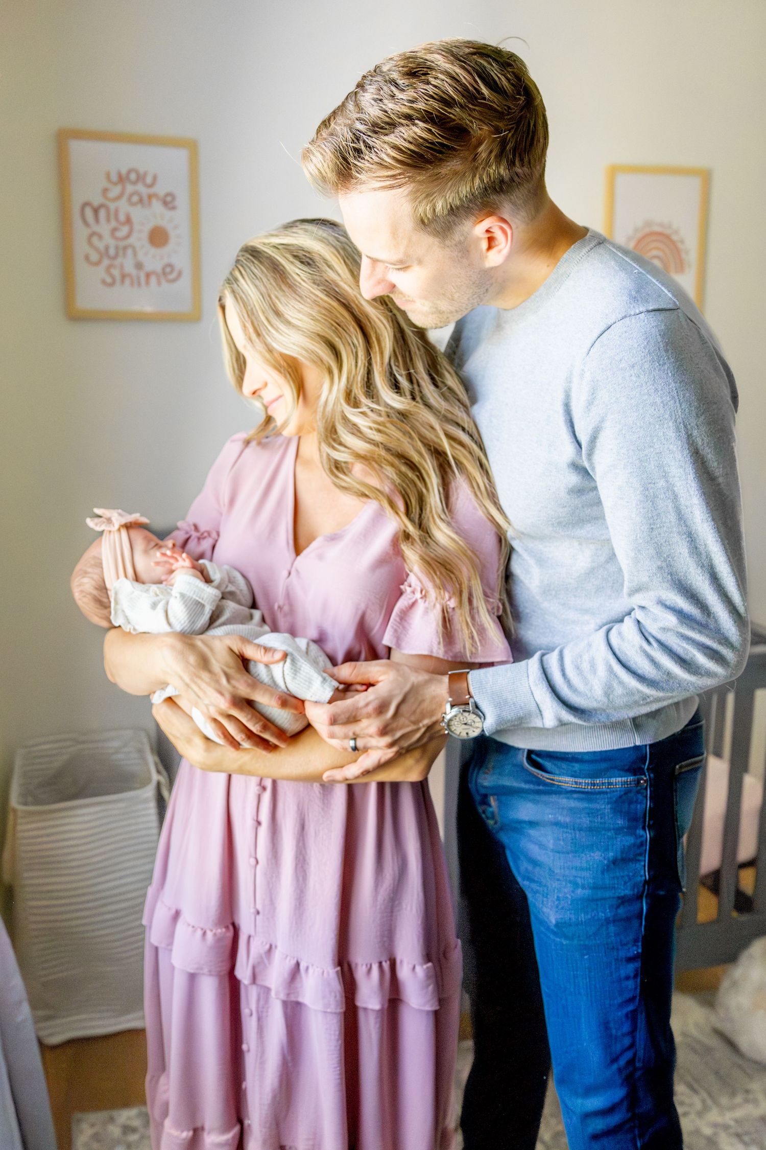 New parents in cozy nursery hold their sleeping newborn while standing near framed wall art.