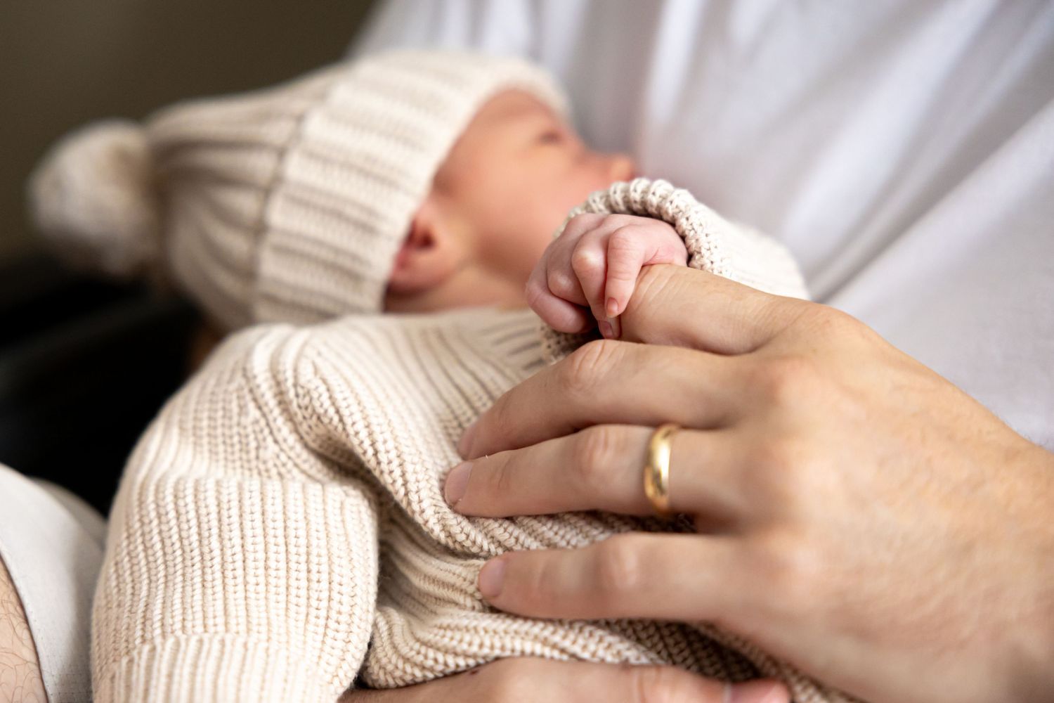 Close-up of tiny newborn hand grasping parent's finger with wedding ring visible.