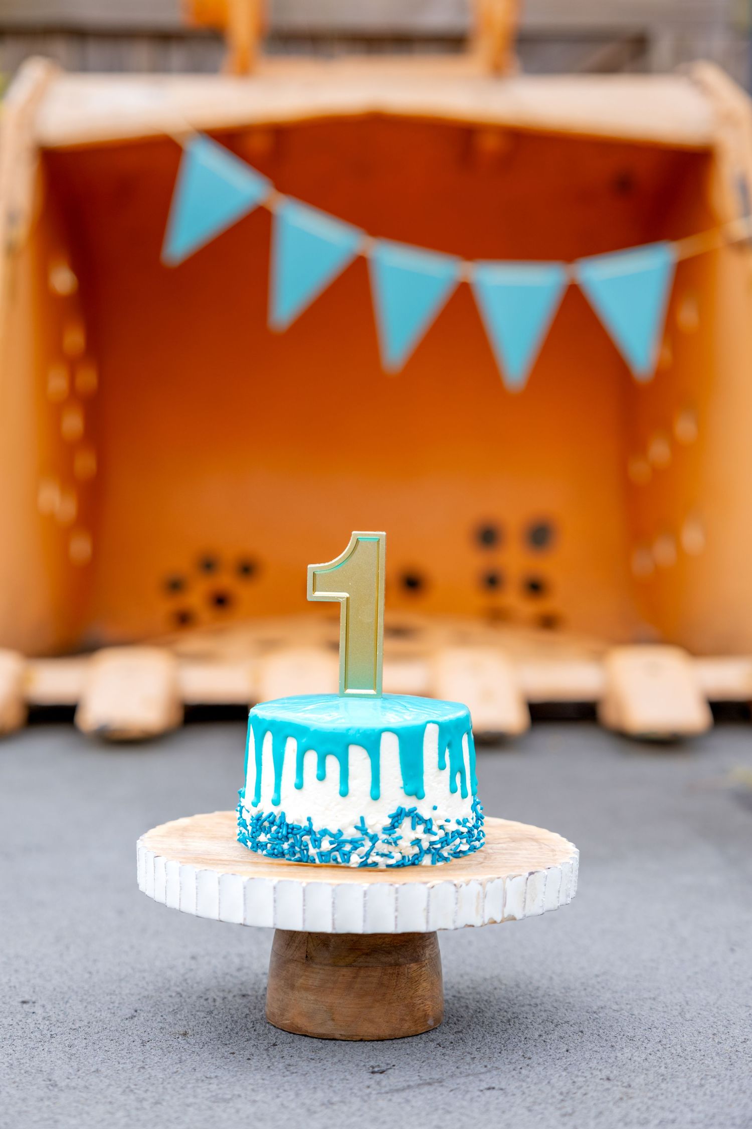 A blue and white smash cake with number 1 topper displayed under turquoise bunting decorations and excavator scoop at Camp North End.