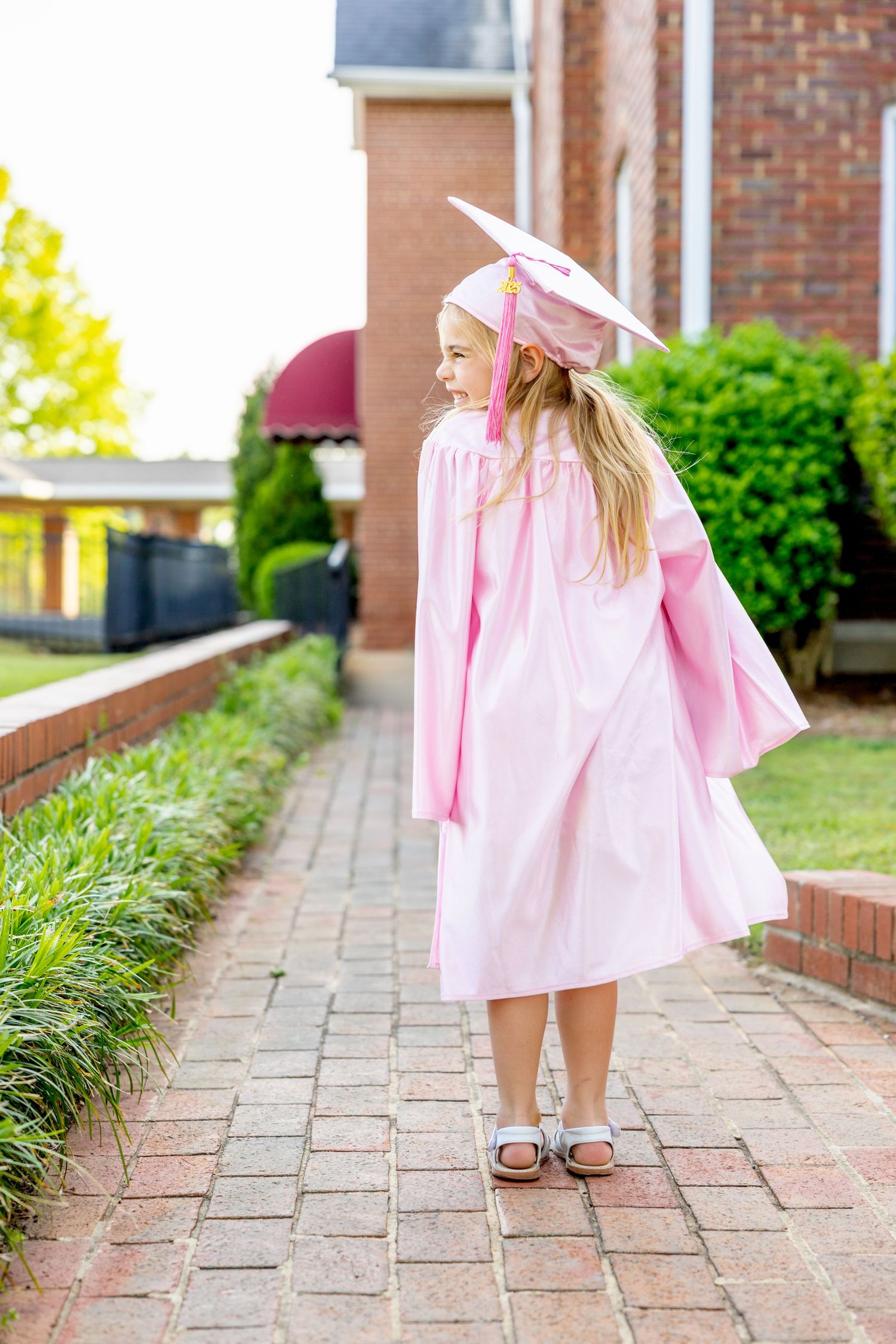 Side view of small preschool graduate walking along brick pathway in pink cap and gown.