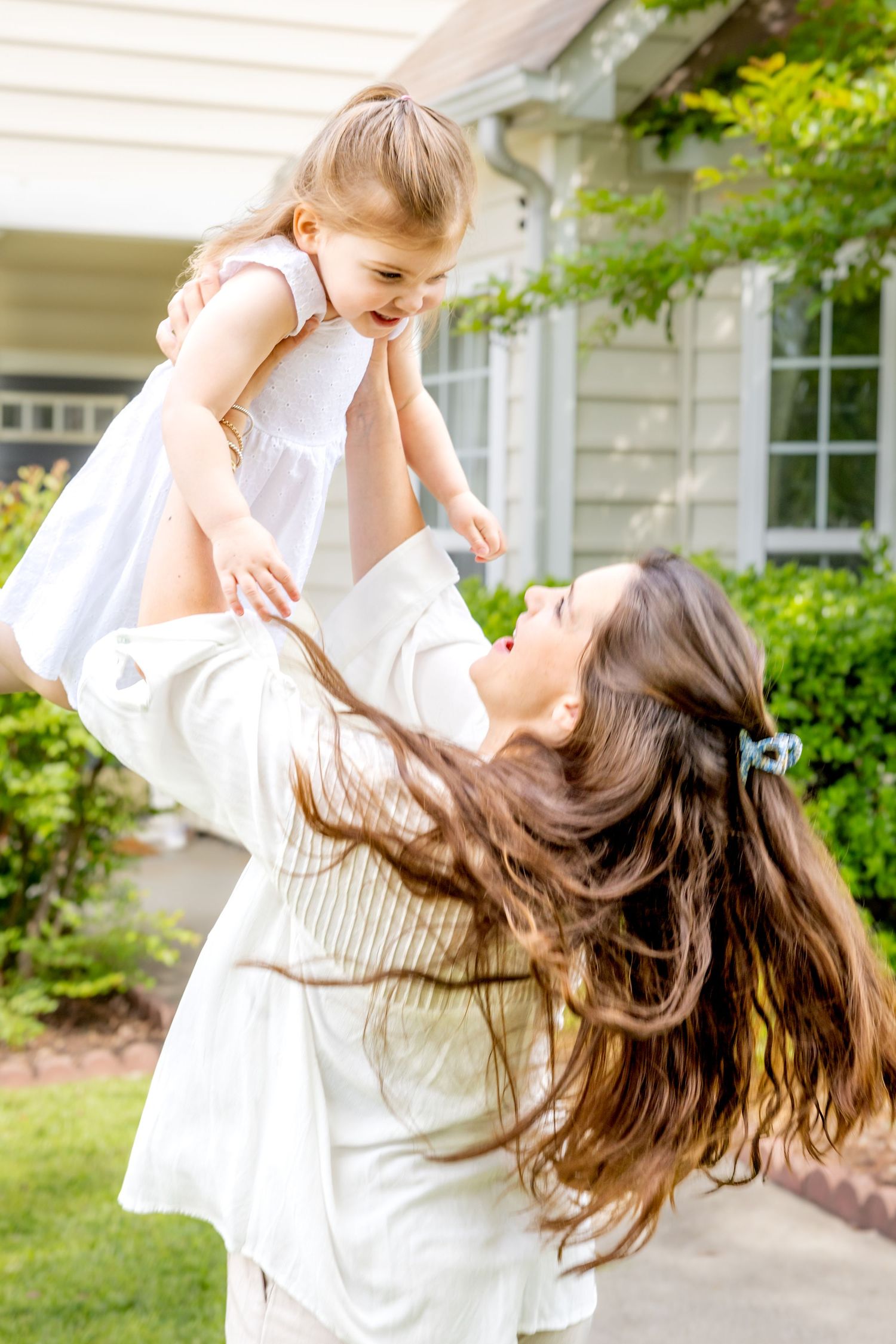 Parent lifting small child in white dress high in the air during playful outdoor moment.