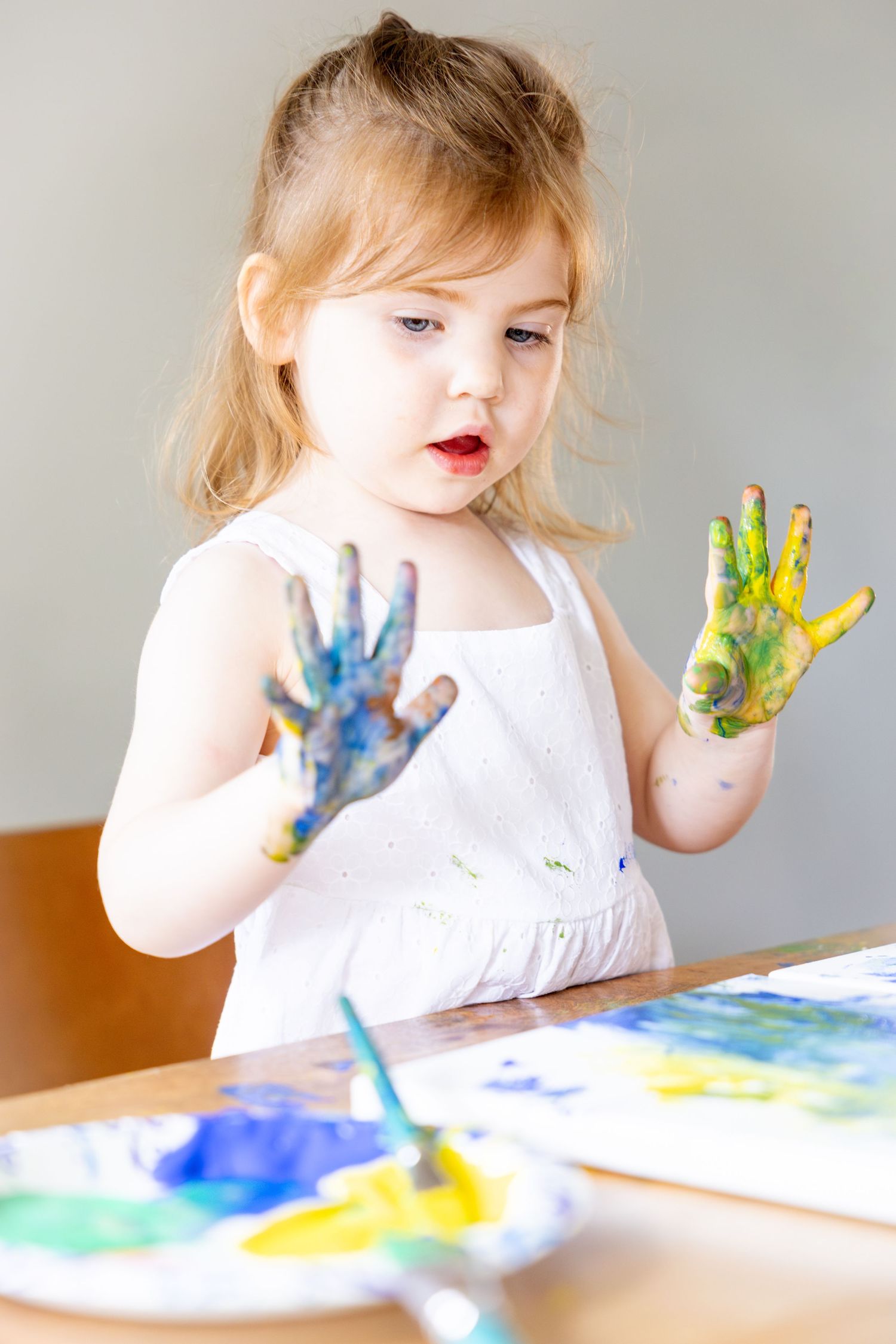Small child in white dress finger painting with blue and yellow paint on hands at a table.