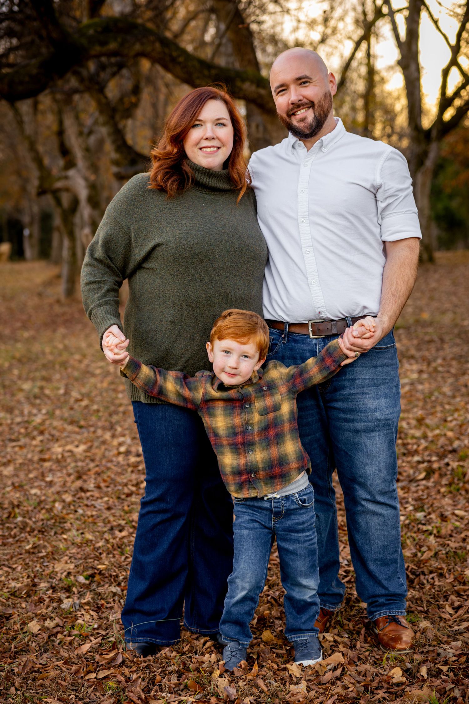 Family of three poses together among fallen autumn leaves in a wooded area at sunset.