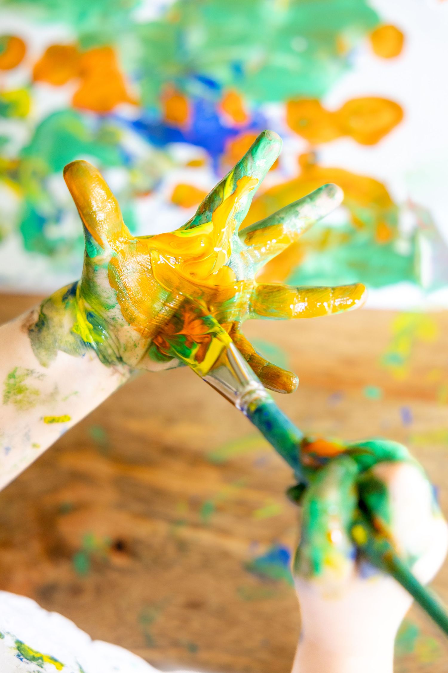 Close up of paint covered hands with yellow and green colors against blurred artwork.