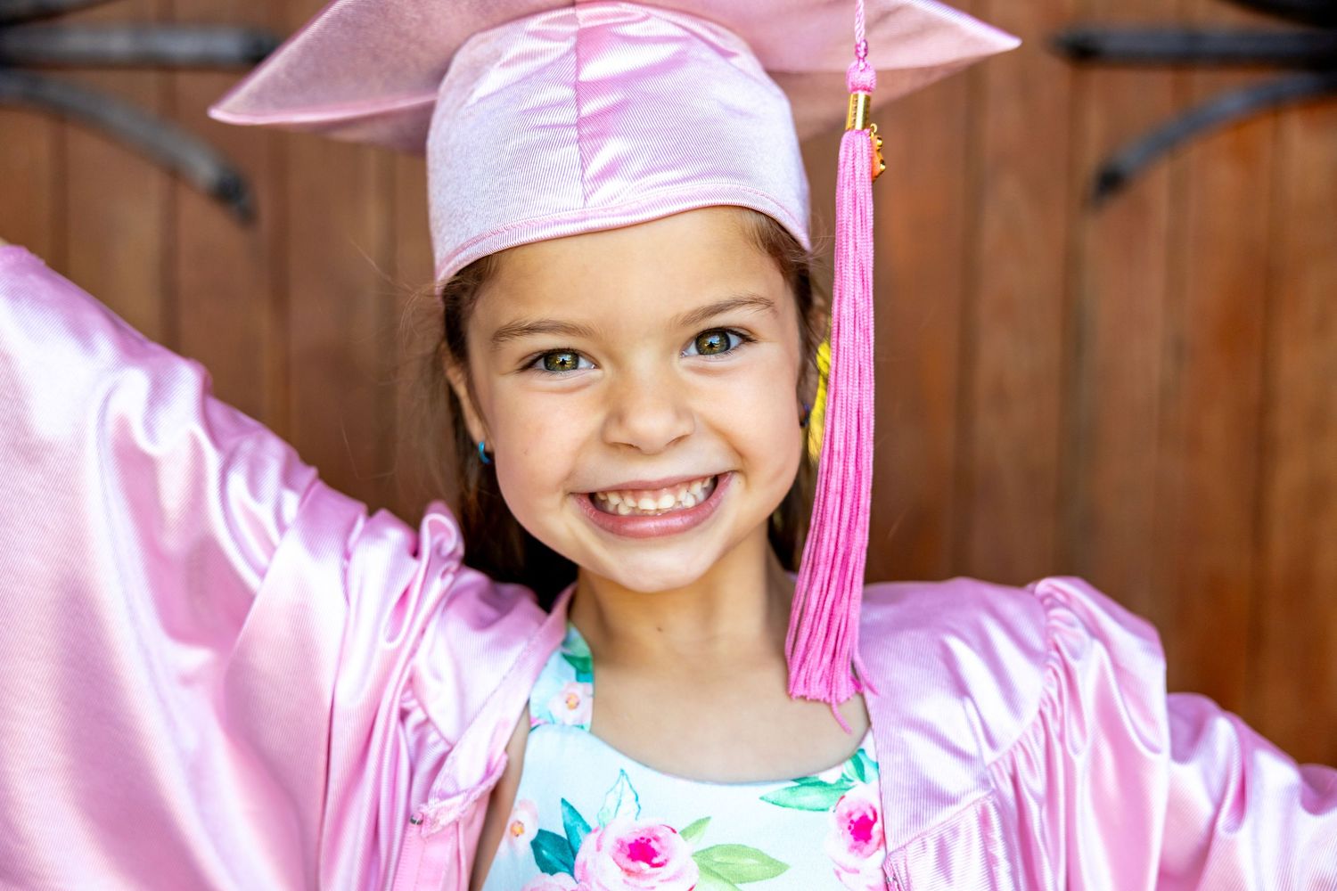Young preschool graduate wearing pink cap and gown smiles brightly against wooden background.