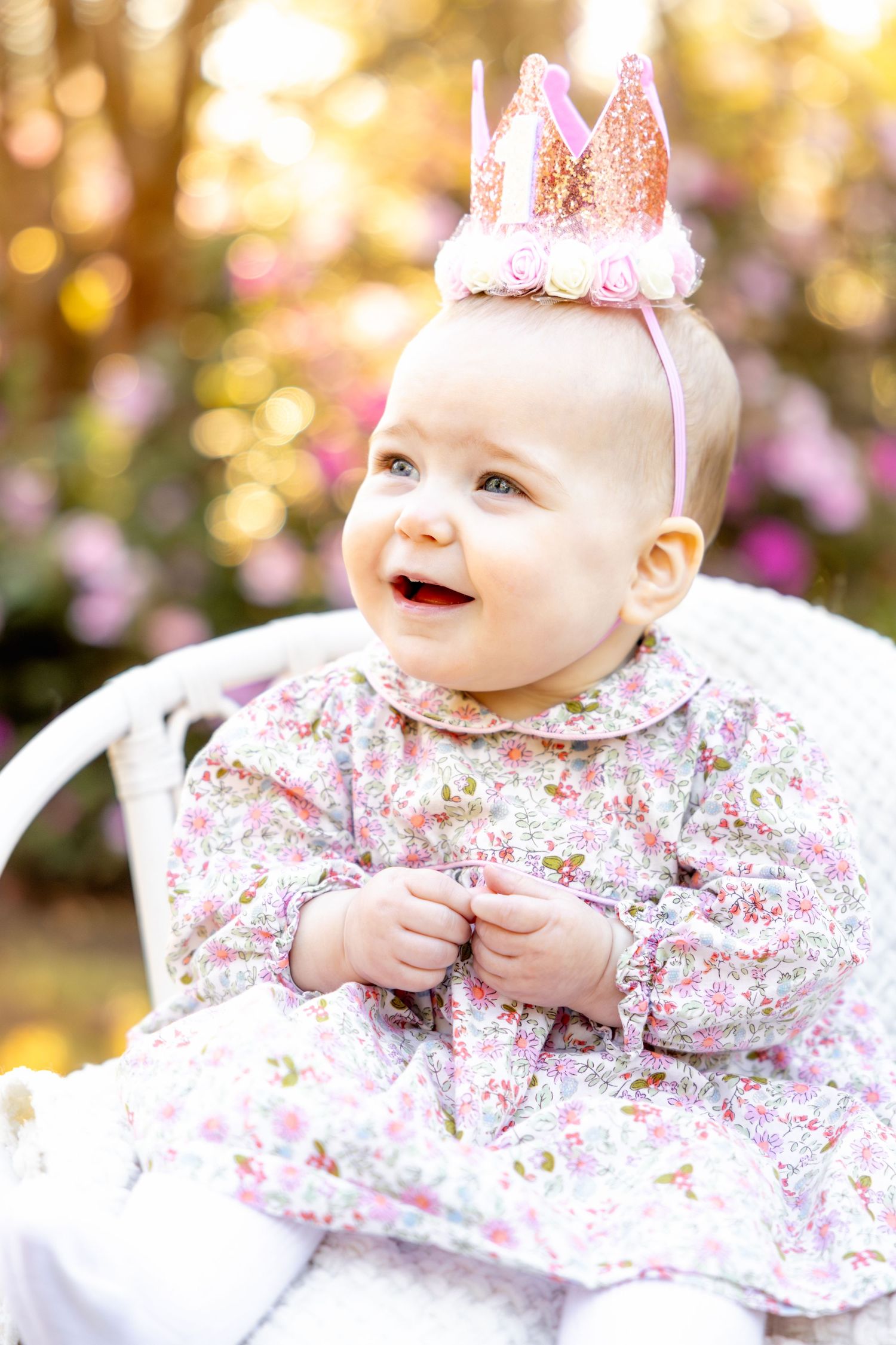 Baby wearing floral dress and pink first birthday crown laughing in sunny garden setting at Glen Cairn Gardens in Rock Hill, SC.