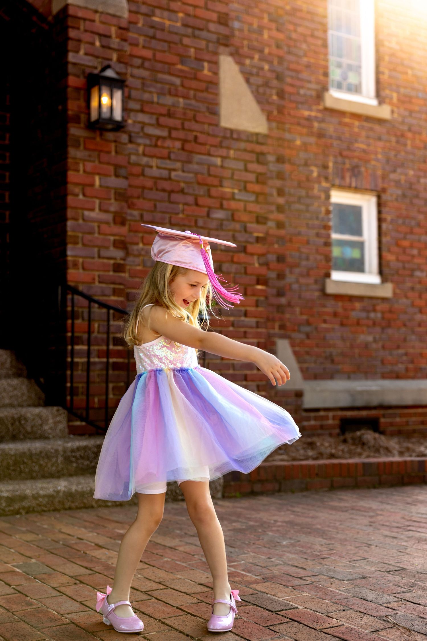 Preschool graduate in pink and purple dress twirls on brick patio near building entrance.