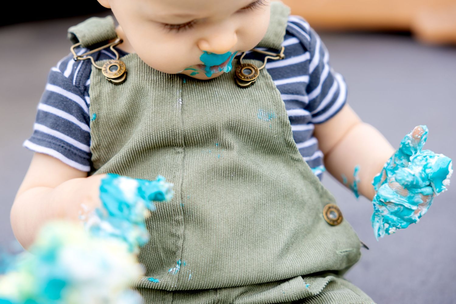 Close-up of green overalls and striped shirt covered in bright blue cake frosting during cake smash session.