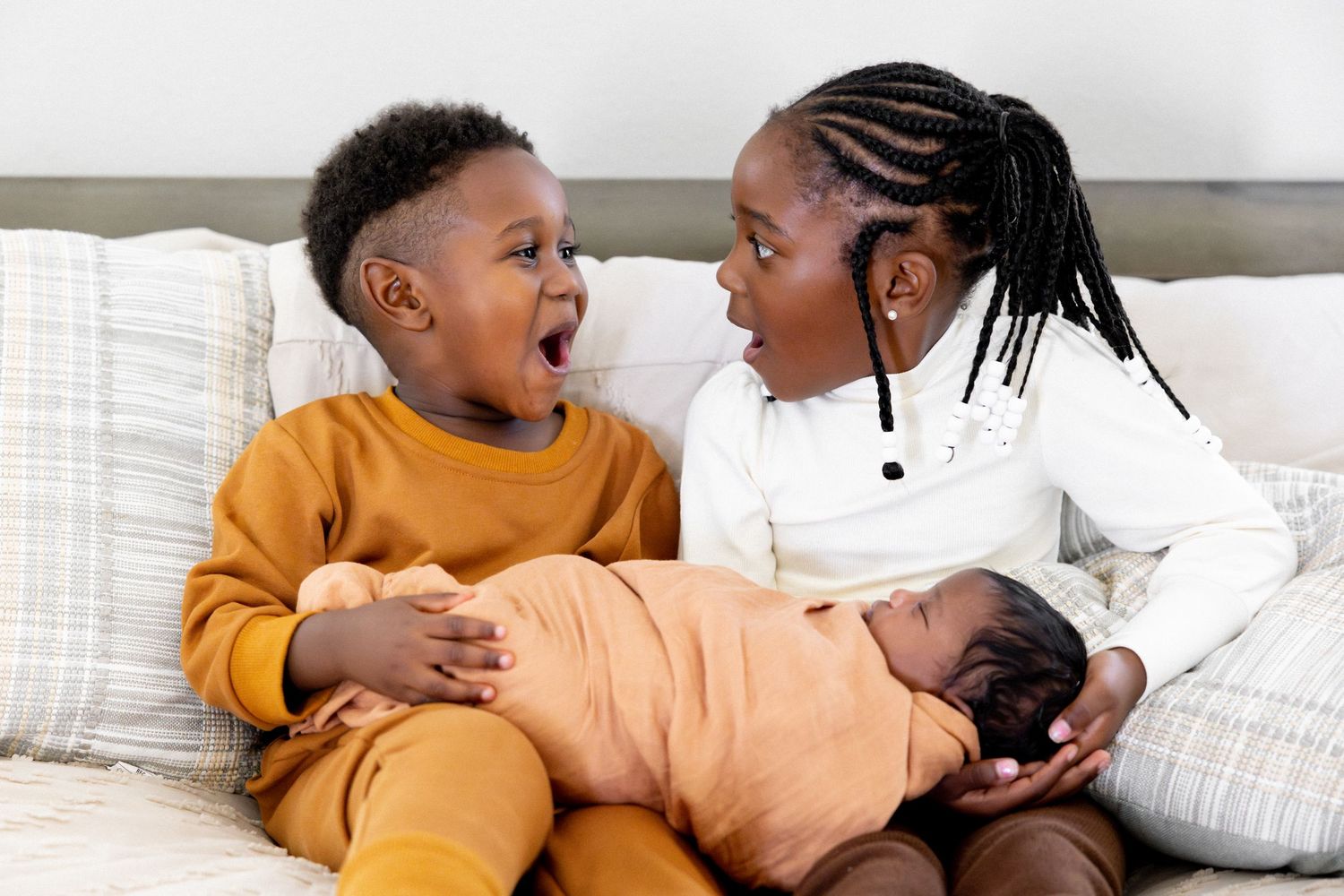 Family sharing joyful moment on white bed with sleeping baby in orange clothing.