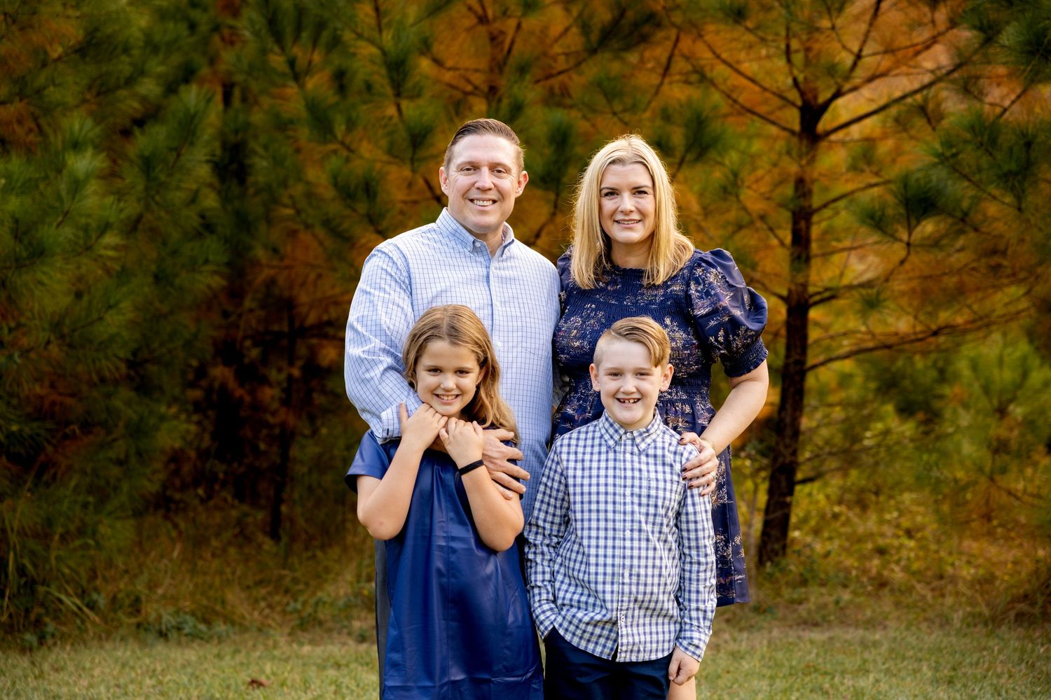 Family portrait with four people standing together against an autumn forest background.