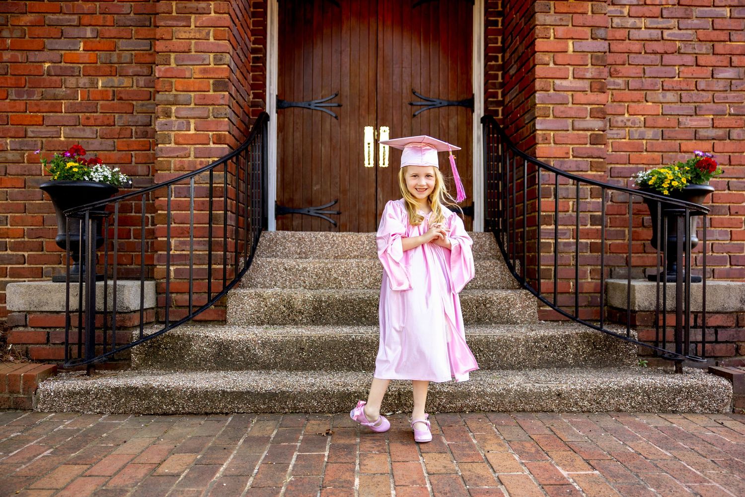 Preschool graduate in pink cap and gown poses on brick steps between planters at entrance.
