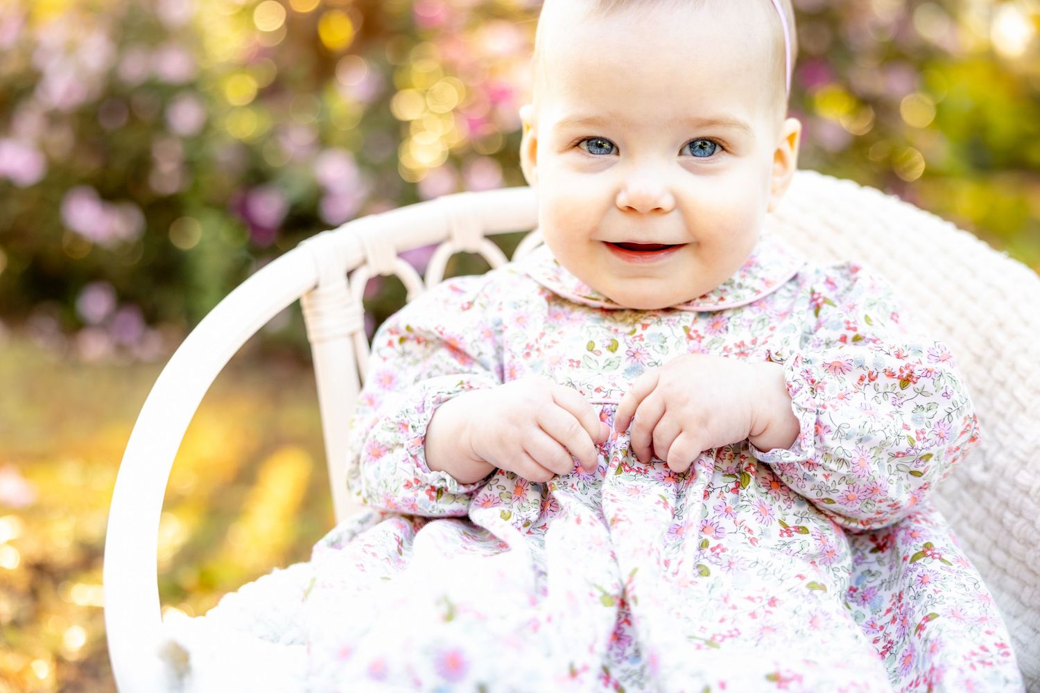 Baby in floral dress with pink first birthday crown smiling in golden sunlit outdoor setting at Glencairn Gardens in Rock Hill, SC.