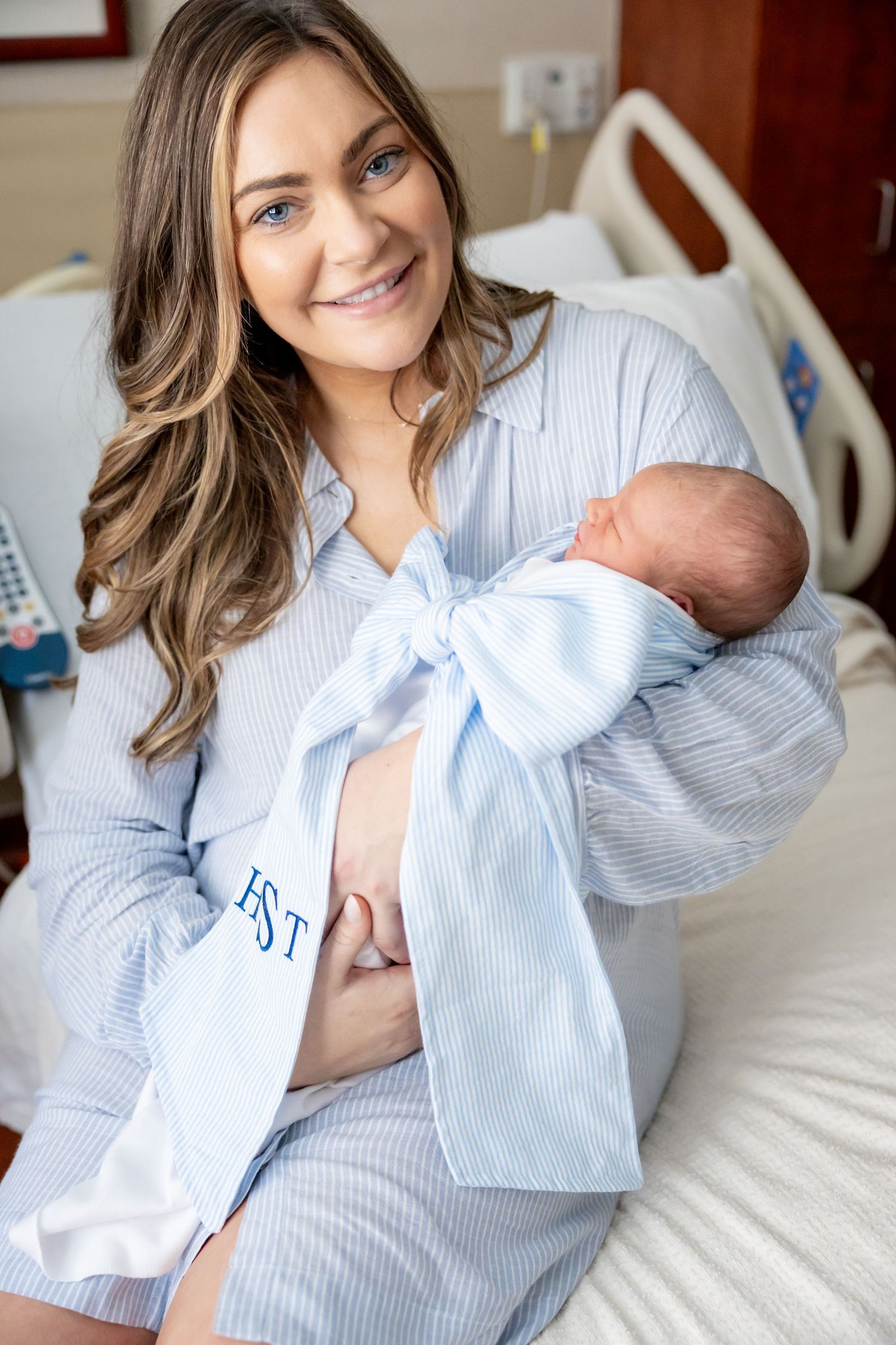 A new mother in a hospital bed holds her sleeping newborn wrapped in blue.