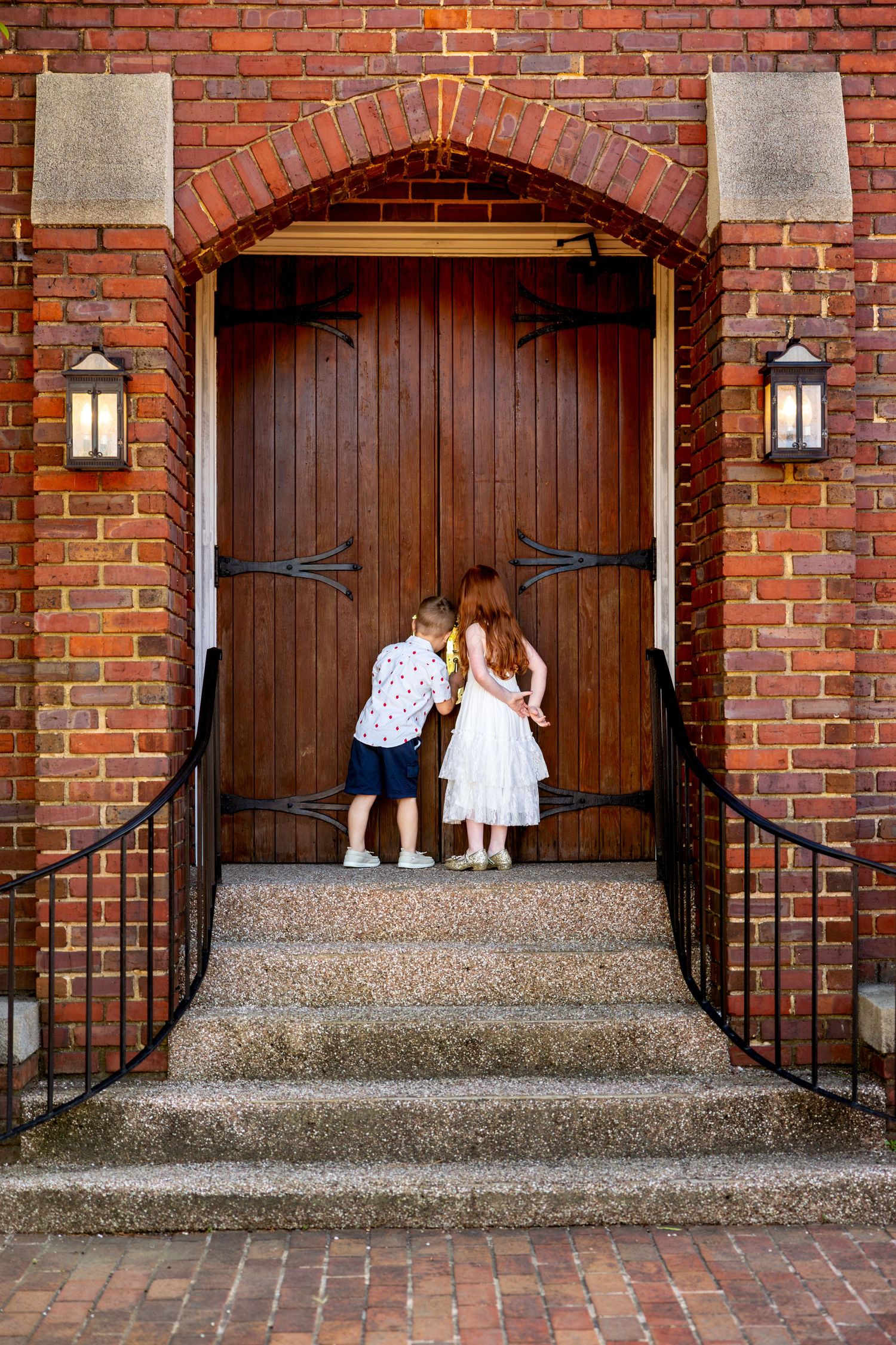 Brick building entrance with wooden doors and steps where children are peeking into door keyhole.