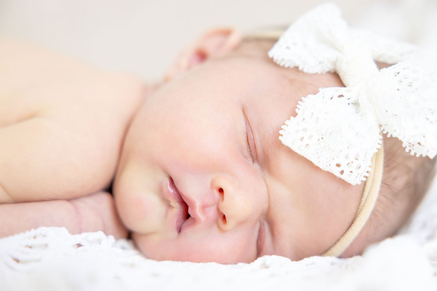Sleeping newborn wearing delicate white lace headband on soft white bedding.