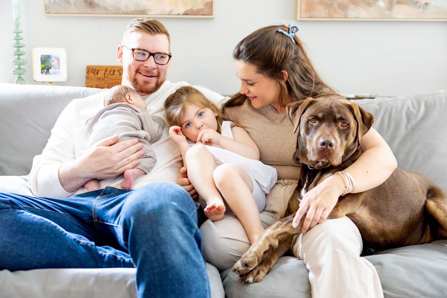 Happy family relaxes on couch with their brown dog while holding baby.