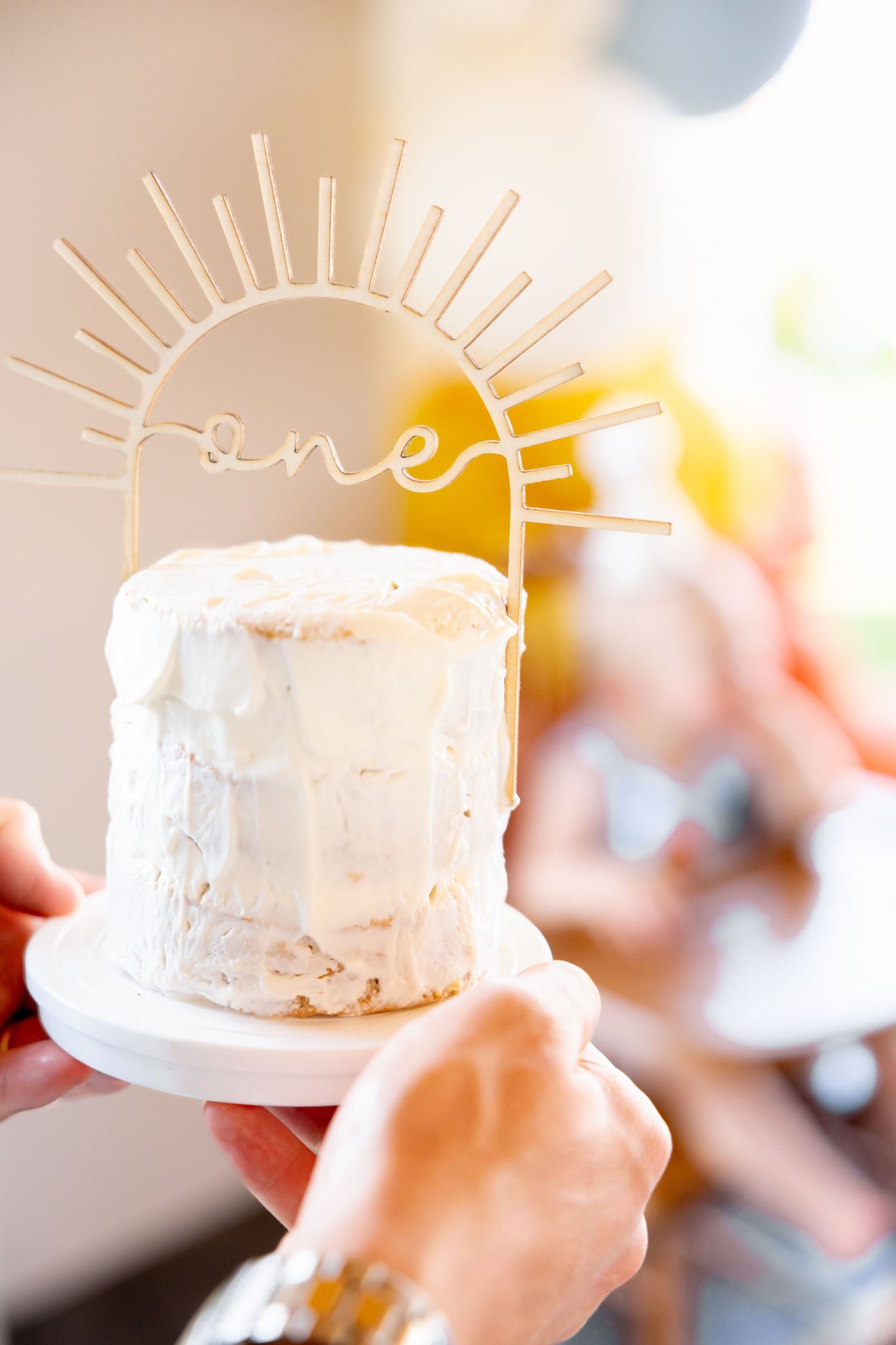 White birthday cake with gold 'one' sun topper being held against balloon backdrop.