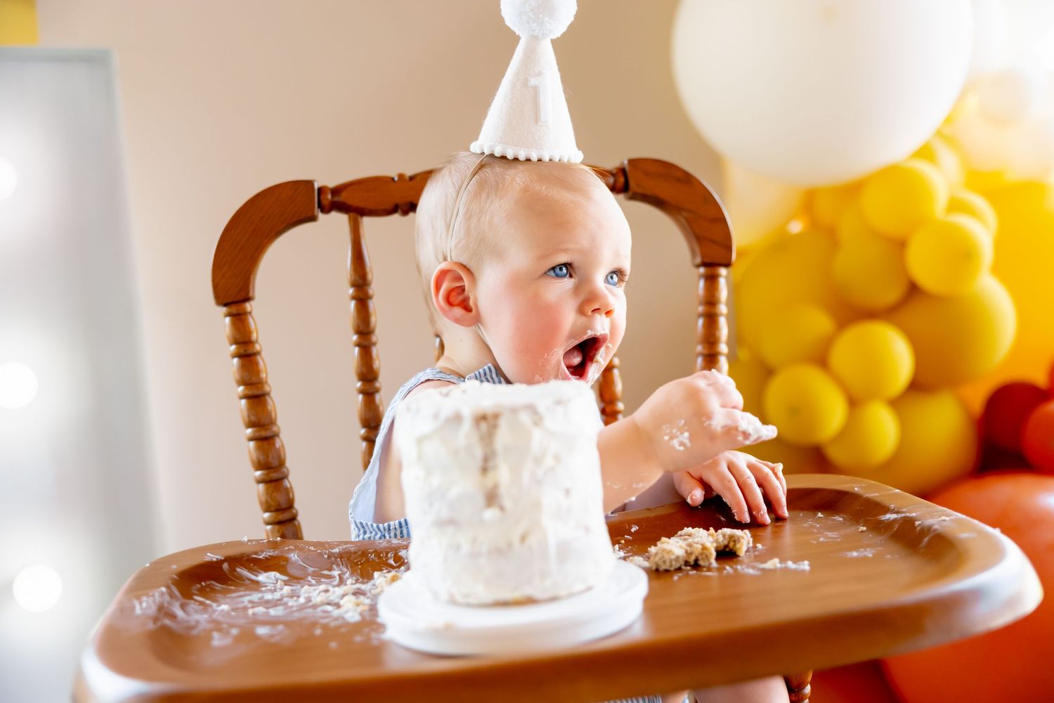 Close up shot of baby in high chair with cake and yellow balloons in background.