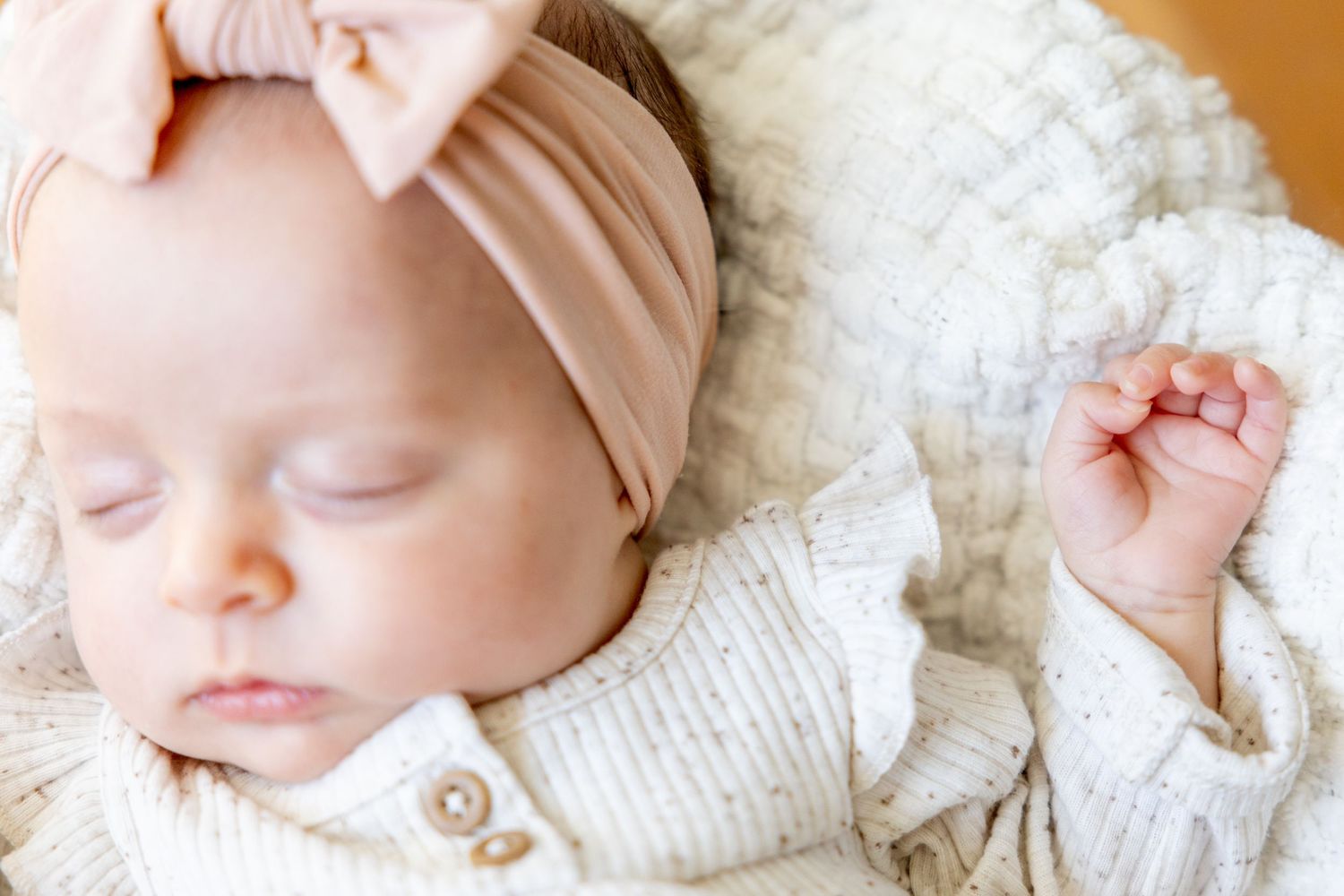 Sleeping baby wearing white knit sweater and pink bow headband rests on soft white blanket.