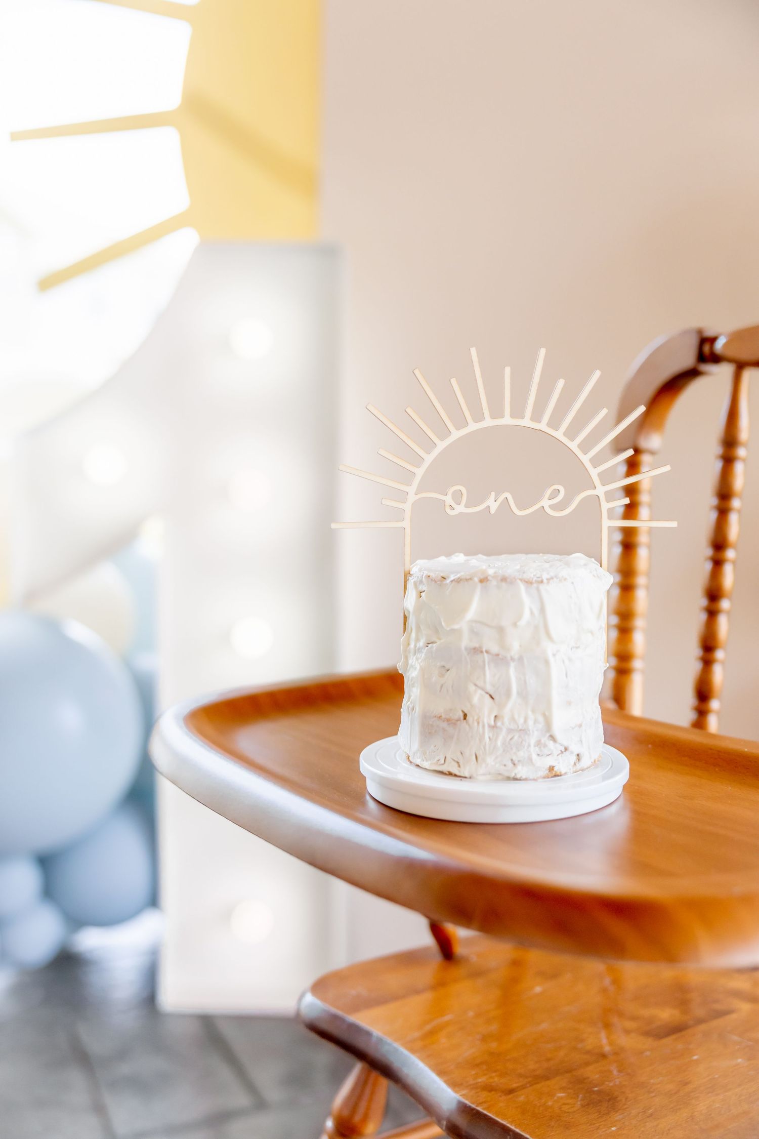 White birthday cake on wooden high chair with decorative sun topper.
