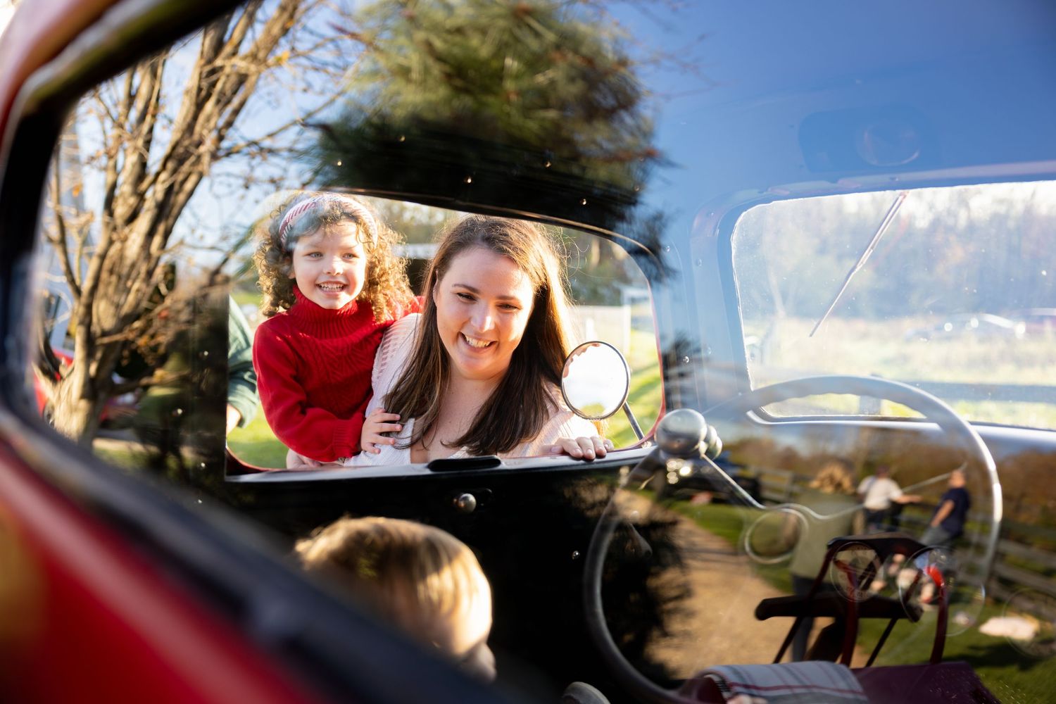 A view through a vintage car's side mirror shows people reflected against a sunny outdoor backdrop.