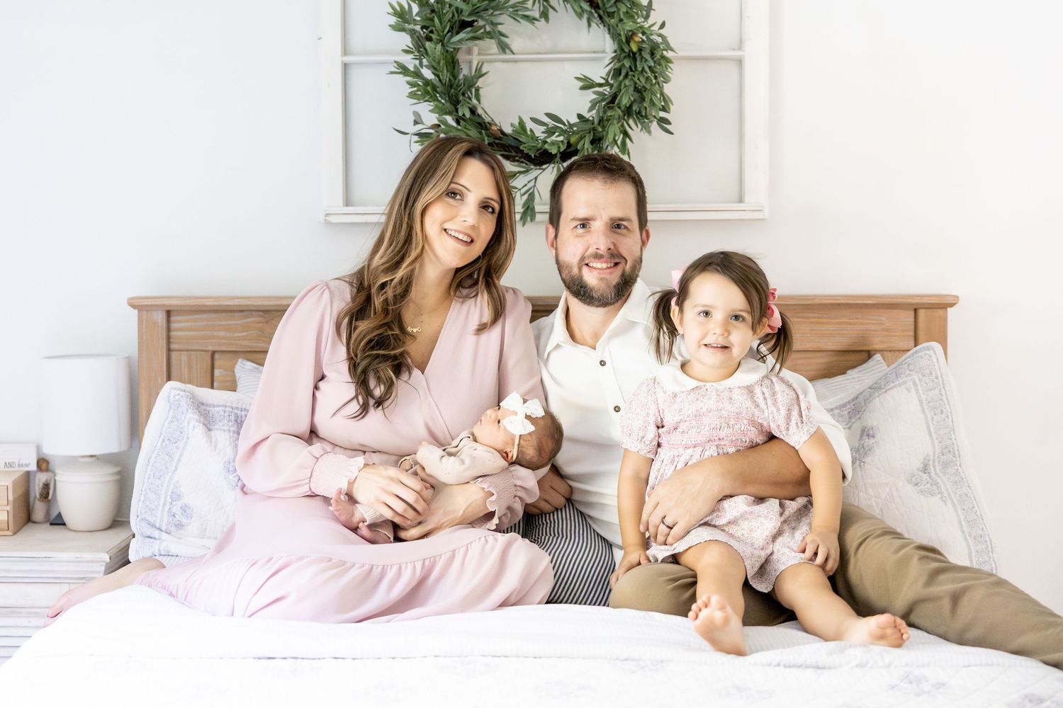 Family relaxing together on white bed with wooden headboard and greenery wreath.