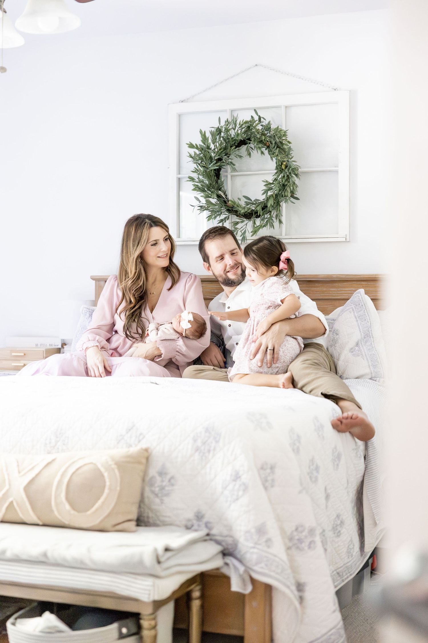 A cozy family moment on a white bed with decorative wreath on the wall during an intimate photoshoot.