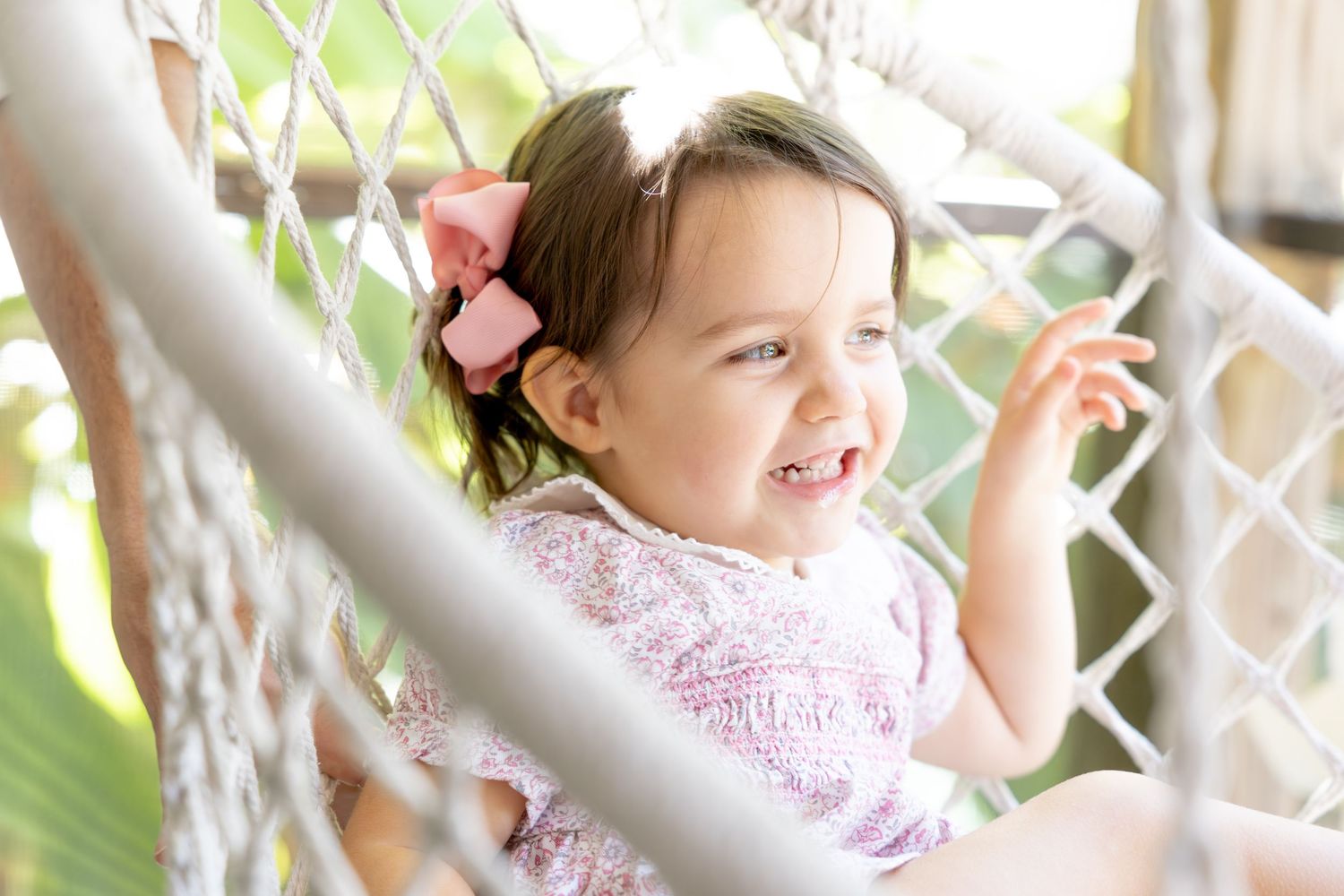 Child sitting in white hammock with pink flower accessory outdoors.