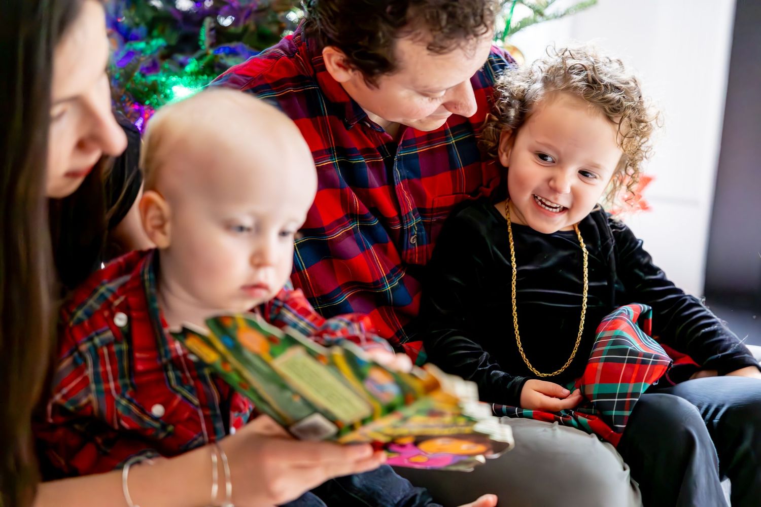 A family reads a Christmas book together while sitting near a decorated tree.