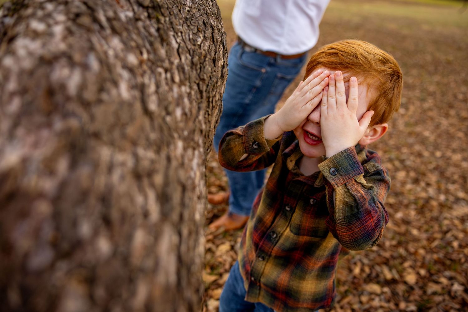 Child playing peek-a-boo behind hands near a large tree trunk.
