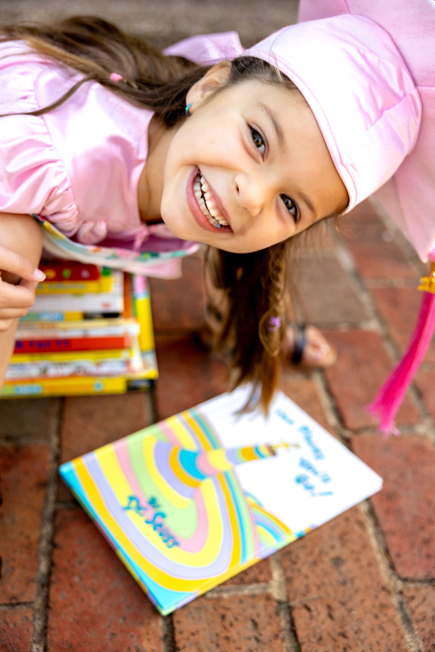 Preschool graduate sitting on stack of books bending down and smiling.