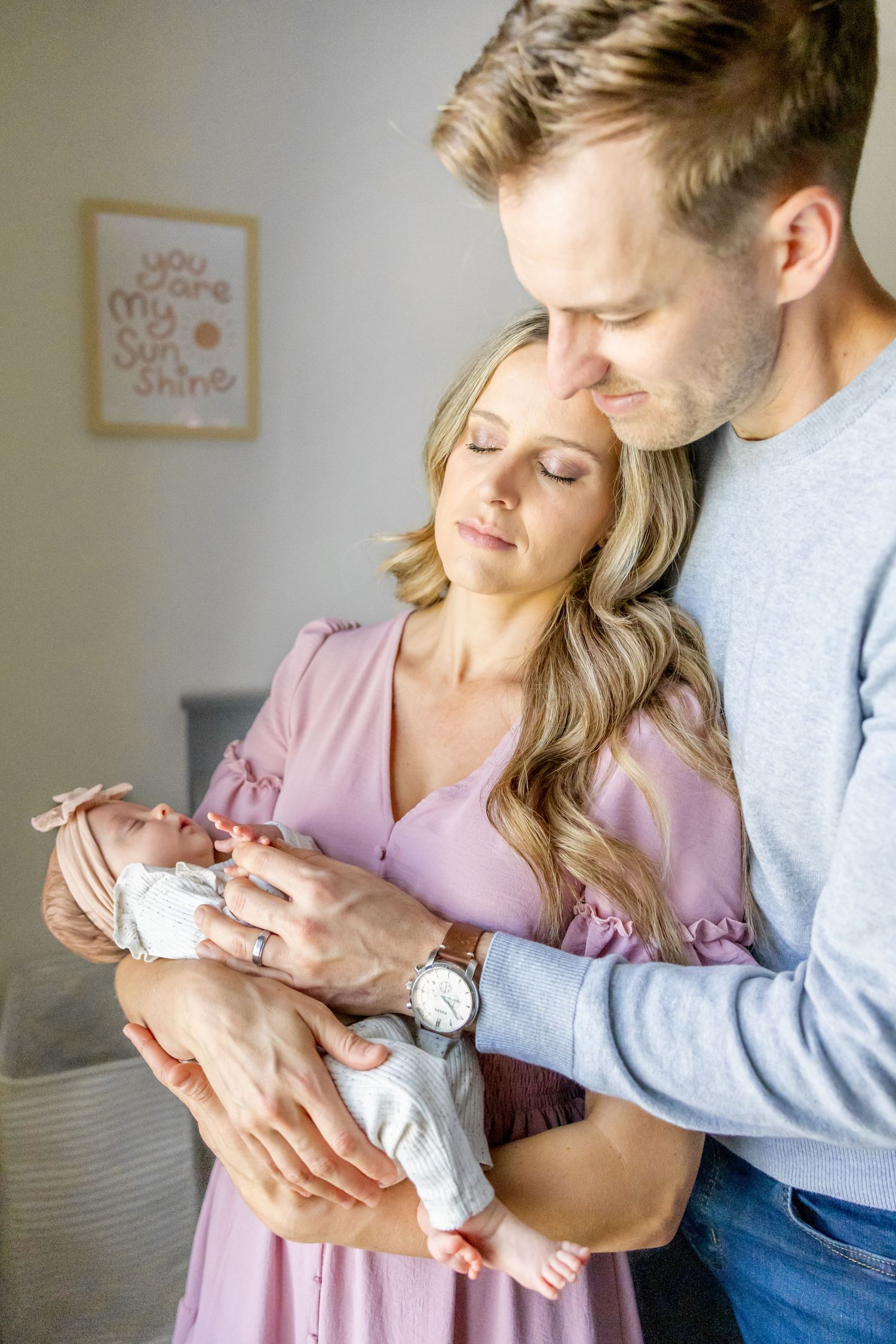 Tender family moment as parents cradle their newborn baby wearing white against a neutral wall background.