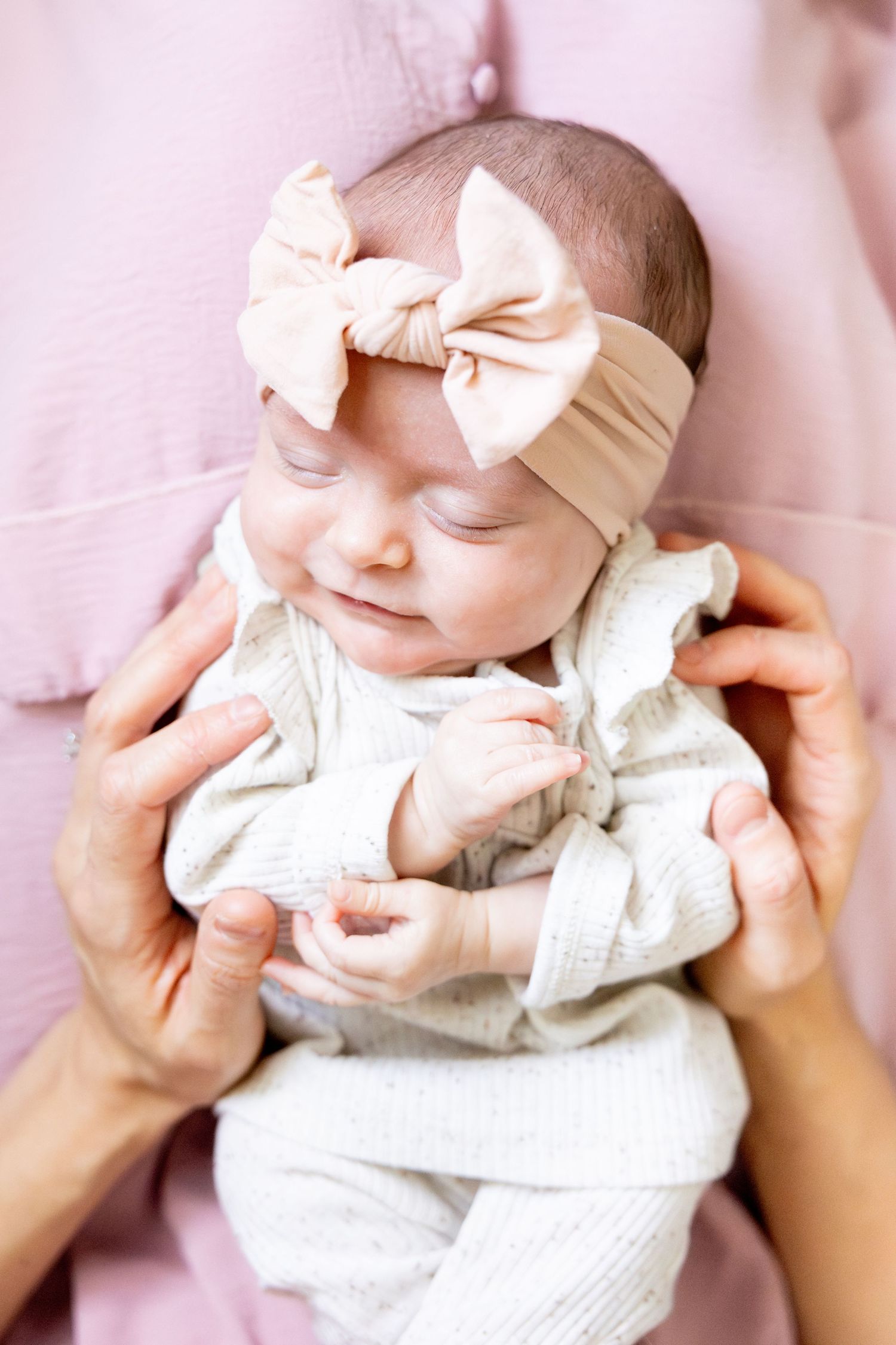 Baby in white ruffled outfit and bow headband being held by hands in pink.