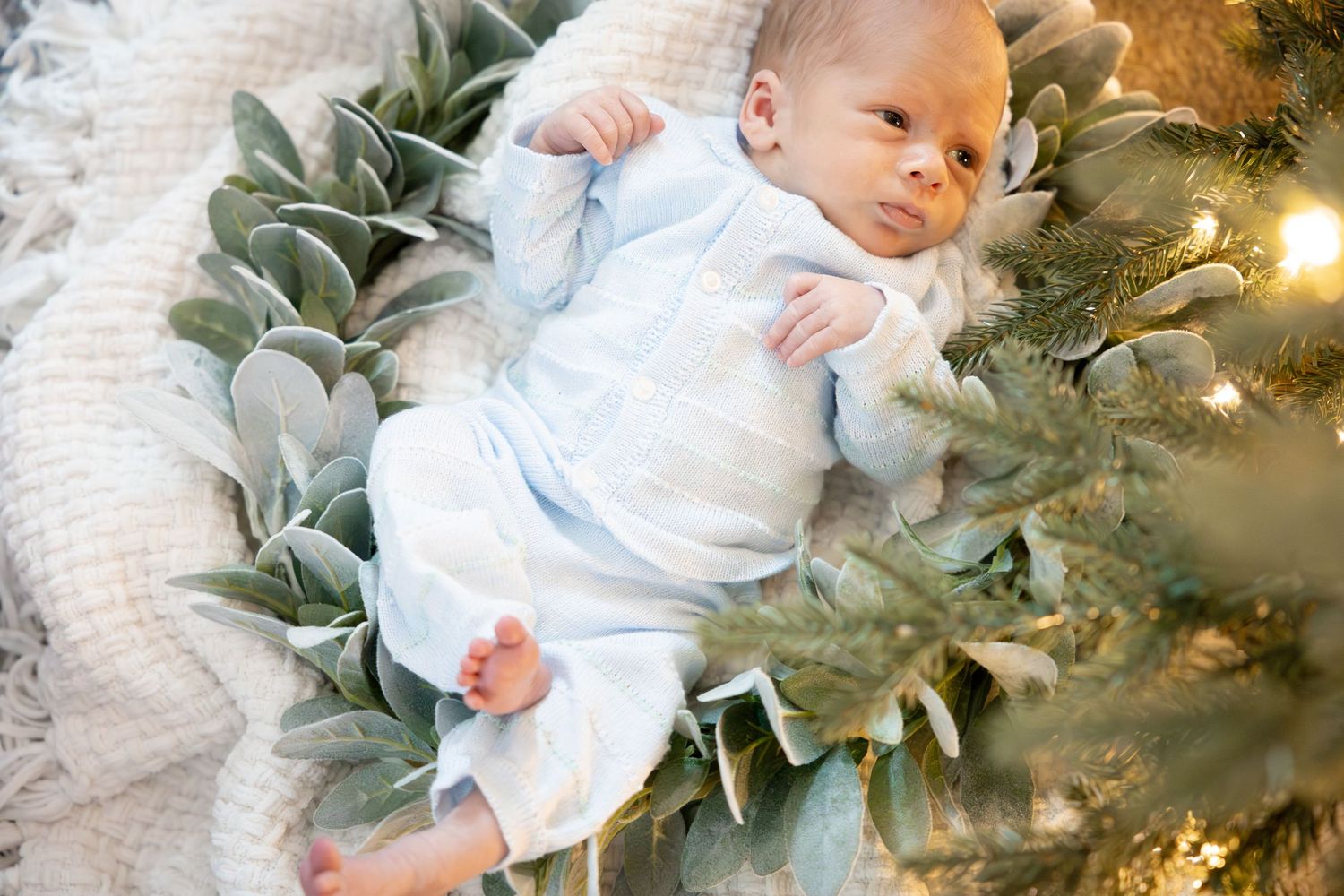 Baby in white outfit lying next to a Christmas tree with holiday lights.