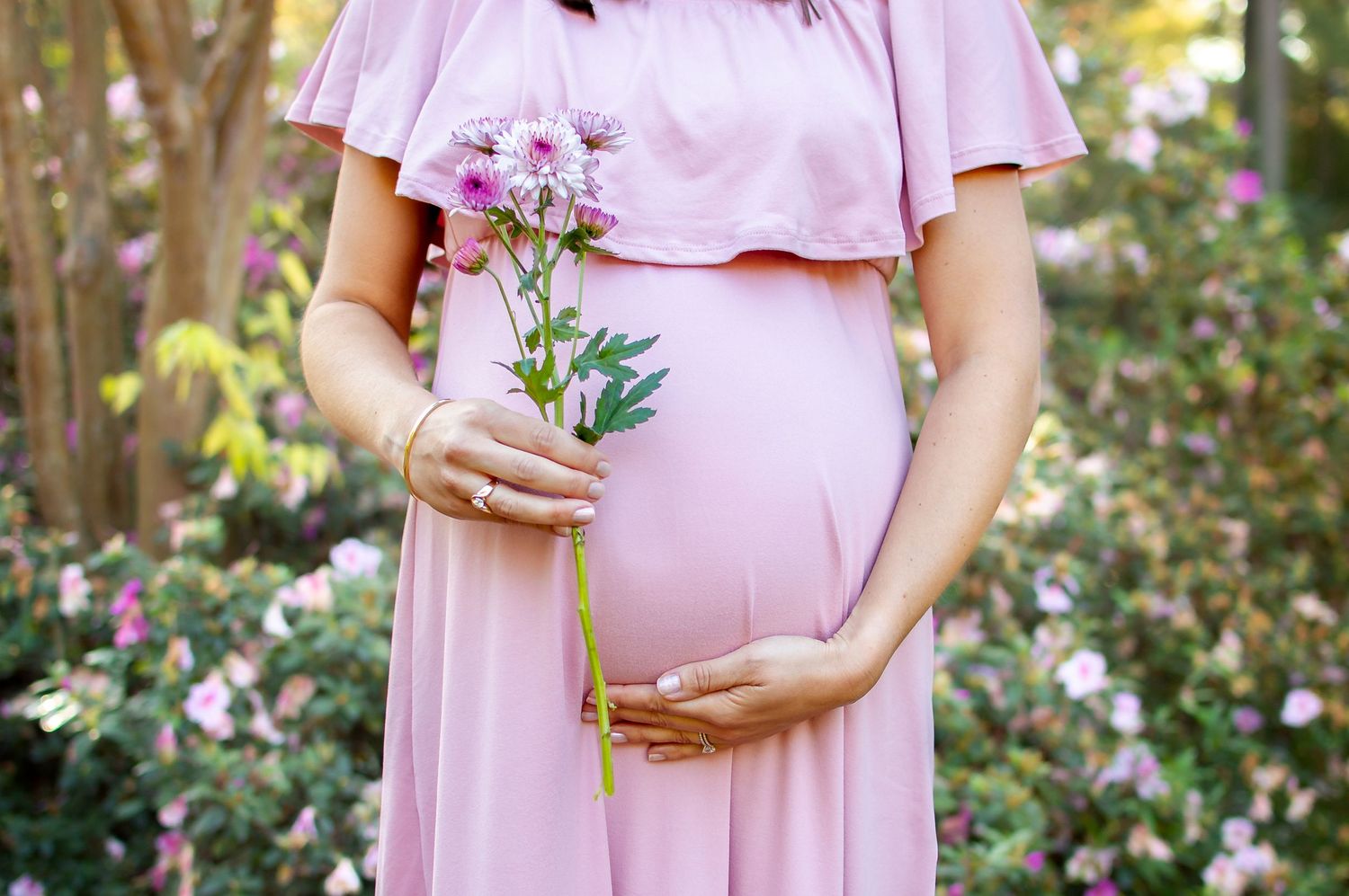 A pregnant woman in pink dress holds pink flowers while cradling her baby bump in a garden.